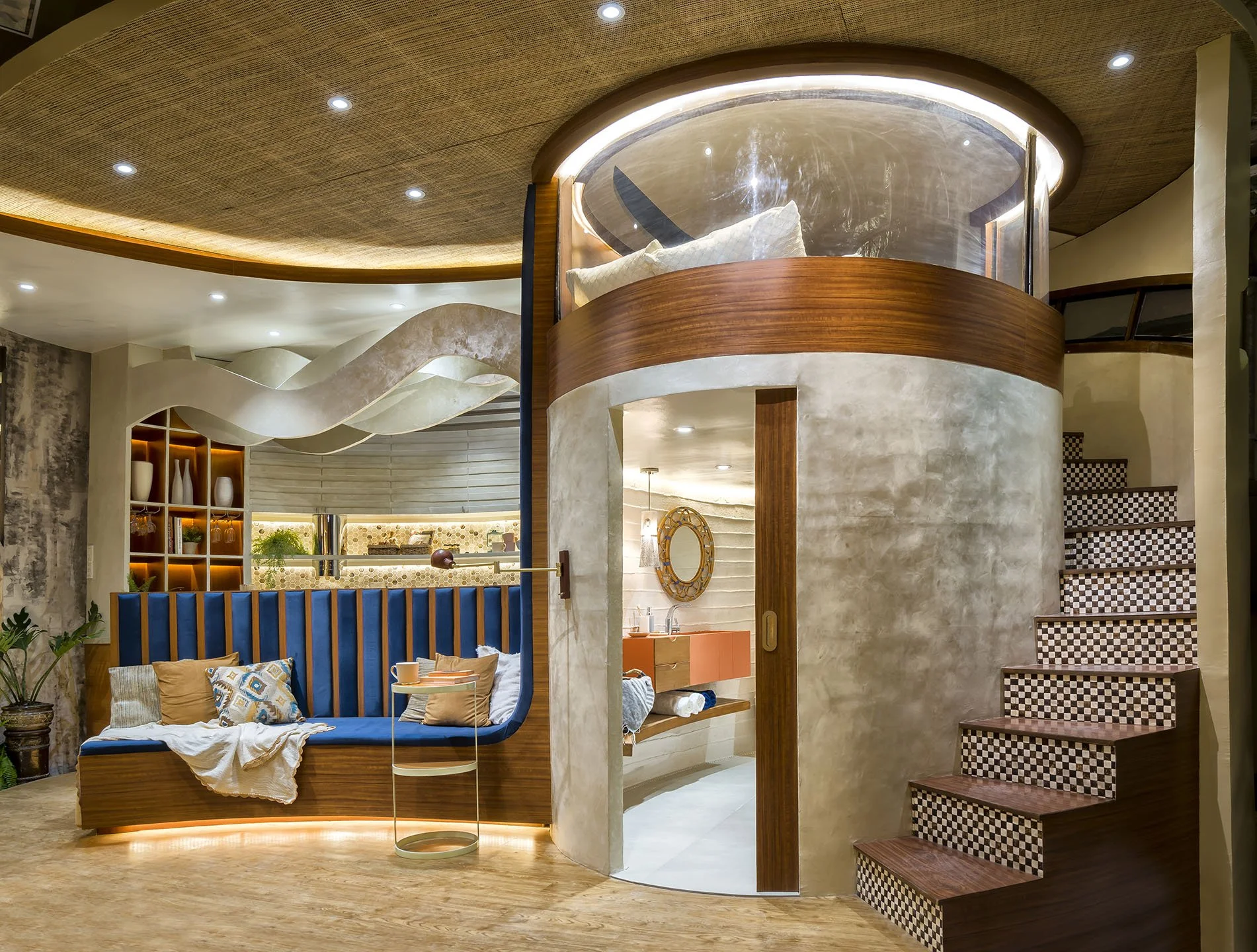 Modern interior space with a curved wooden and glass enclosed bedroom area above a living room. The living room has a sofa with beige pillows and a throw blanket, a small side table, and decorative items.