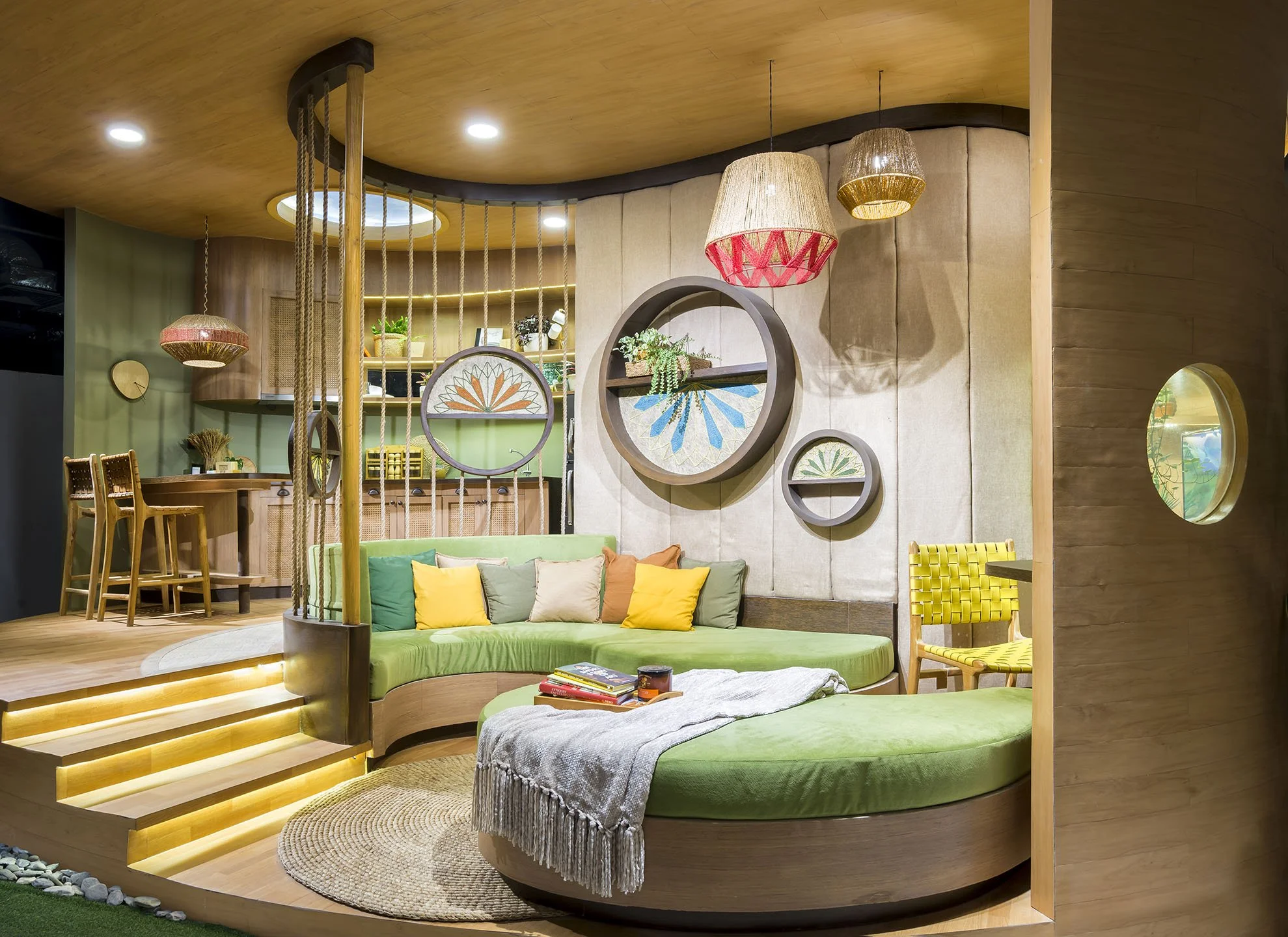 Interior of a modern living space featuring wooden ceiling and floor, green circular seating with colorful pillows, hanging wicker lamps, and decorative wall art with circular frames.