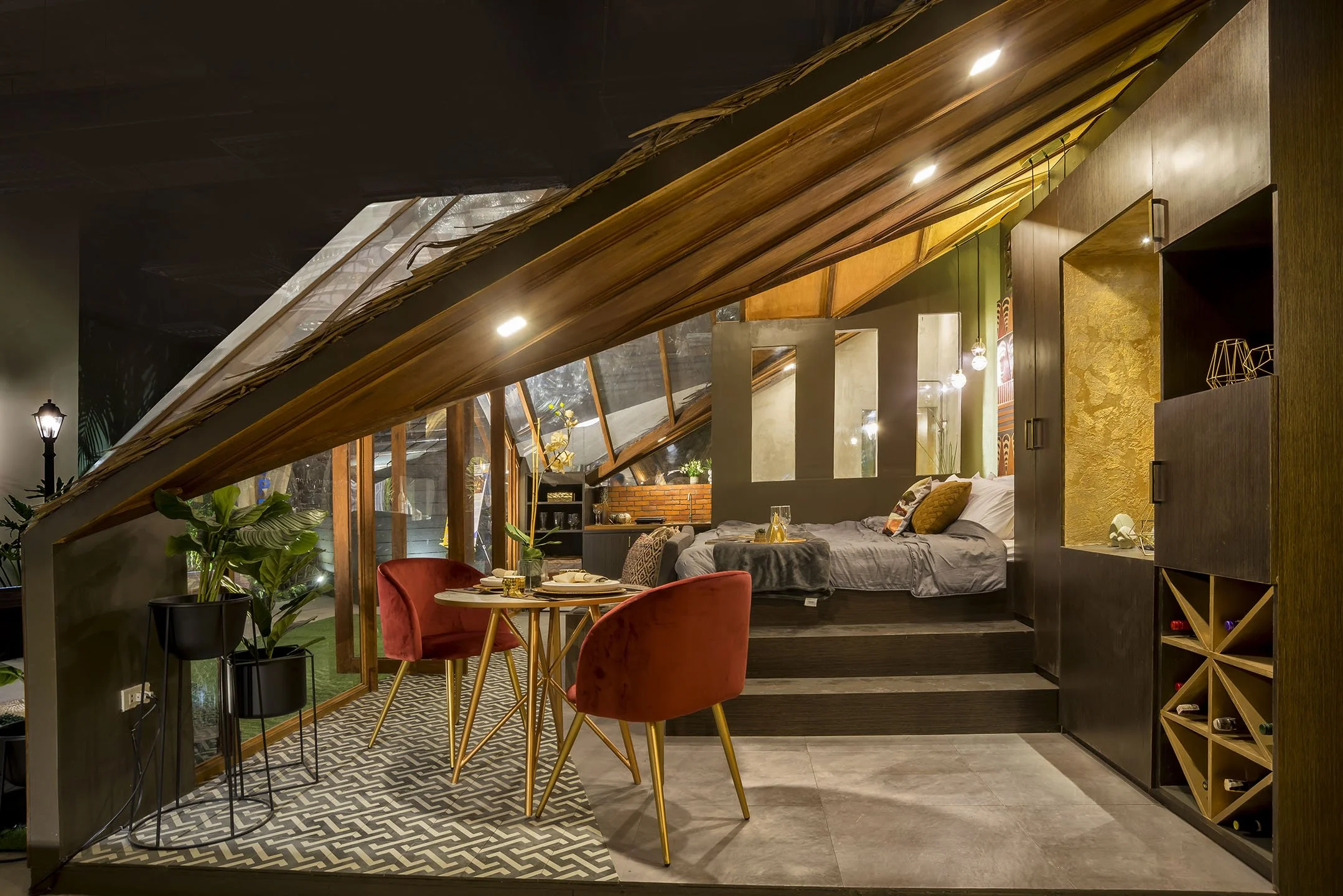 Interior of a modern bedroom with a sloped wooden ceiling, a cozy bed, and a dining area with red velvet chairs, plants, and decorative lighting.
