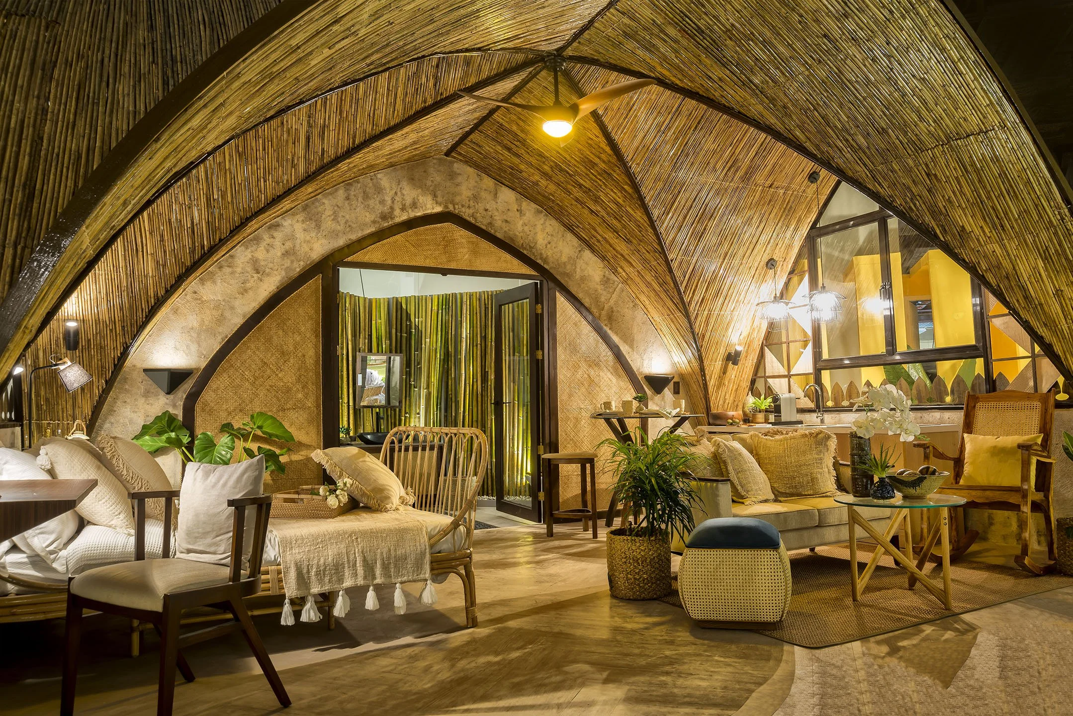 Interior of a room with a bamboo curved ceiling, beige sofa, wooden chairs, and plenty of green plants, decorated in a natural, tropical style with warm lighting.