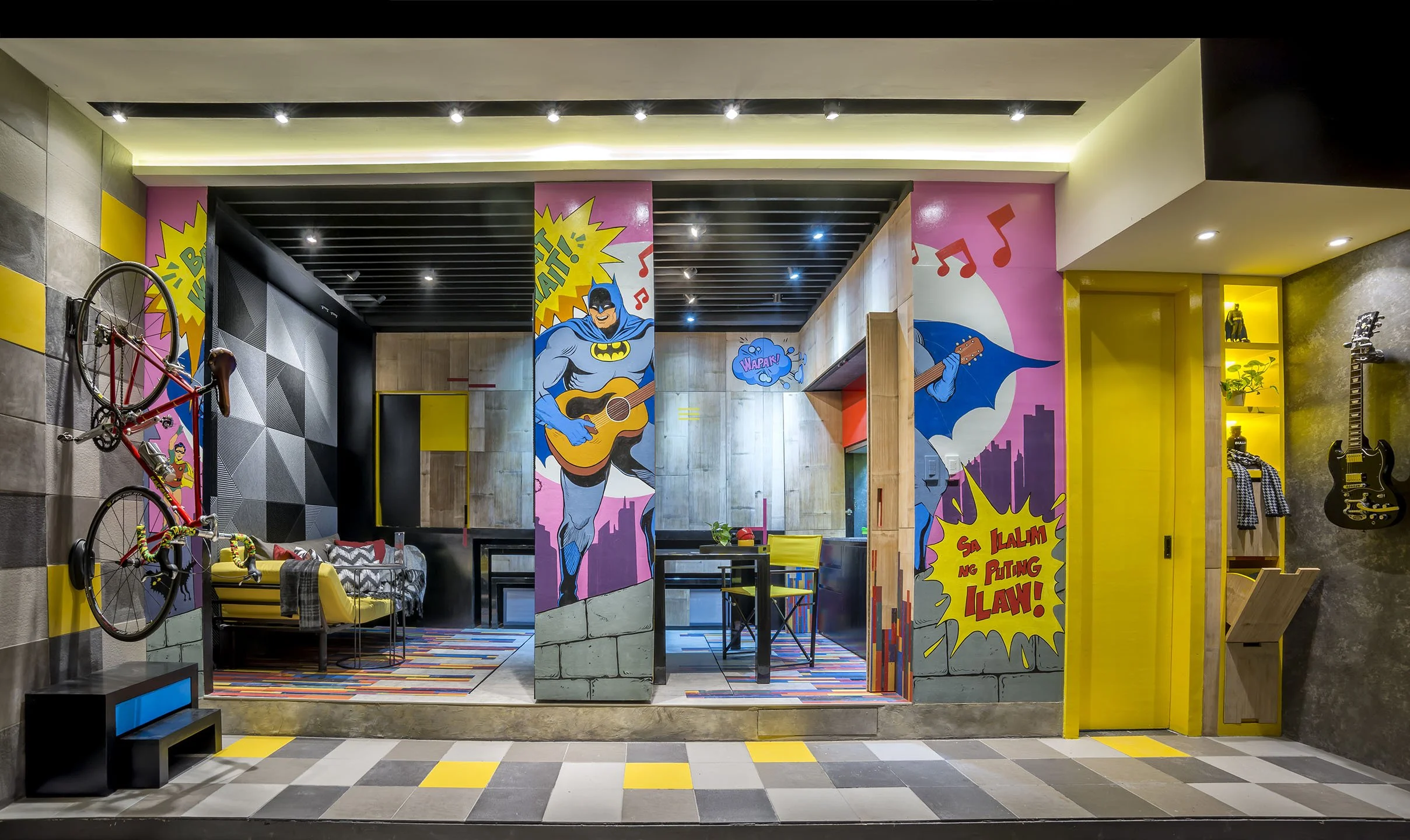Colorful interior with comic-style Batman murals, yellow walls, guitars, a bicycle, and various furniture in a modern, playful design.
