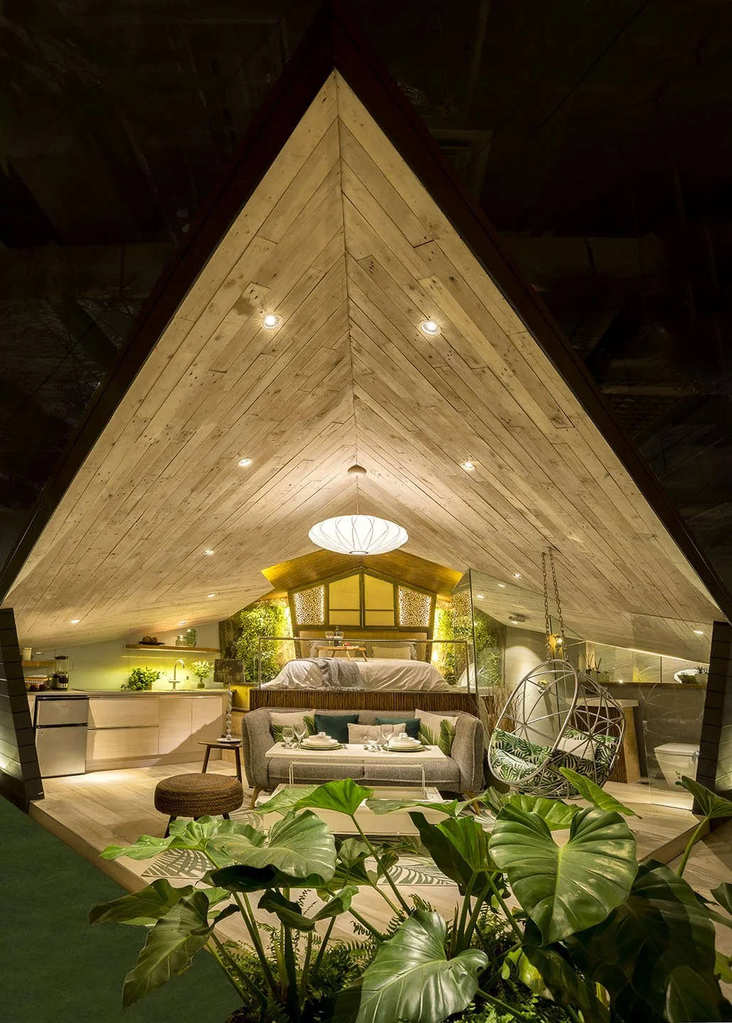 Interior view of an attic style bedroom with wood-paneled ceiling, cozy seating, greenery, and modern decor.