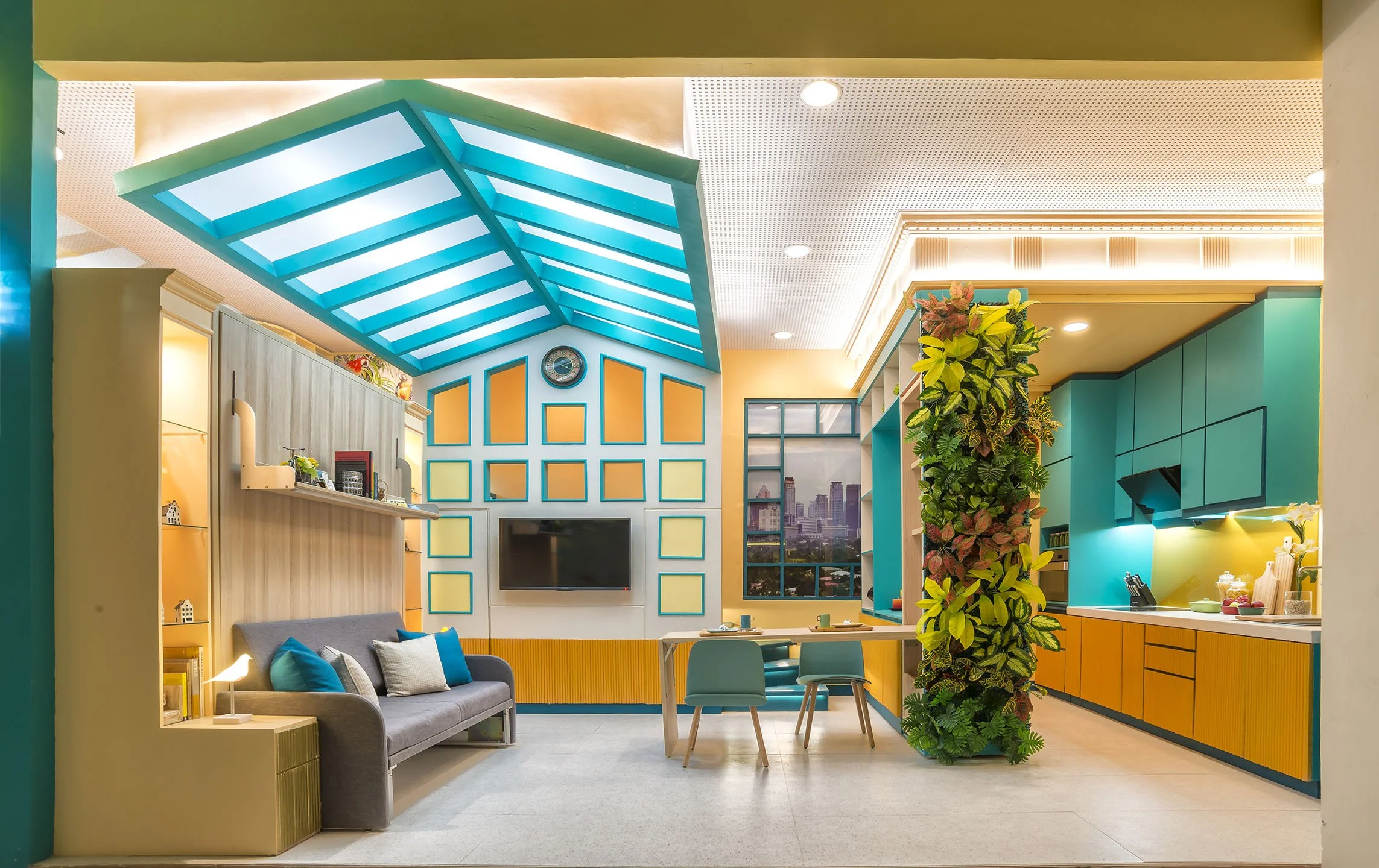 Colorful modern living space with a blue ceiling, yellow accents, and a large plant. Includes a gray sofa with pillows, a TV, a dining table with teal chairs, and a kitchen area with teal cabinets.