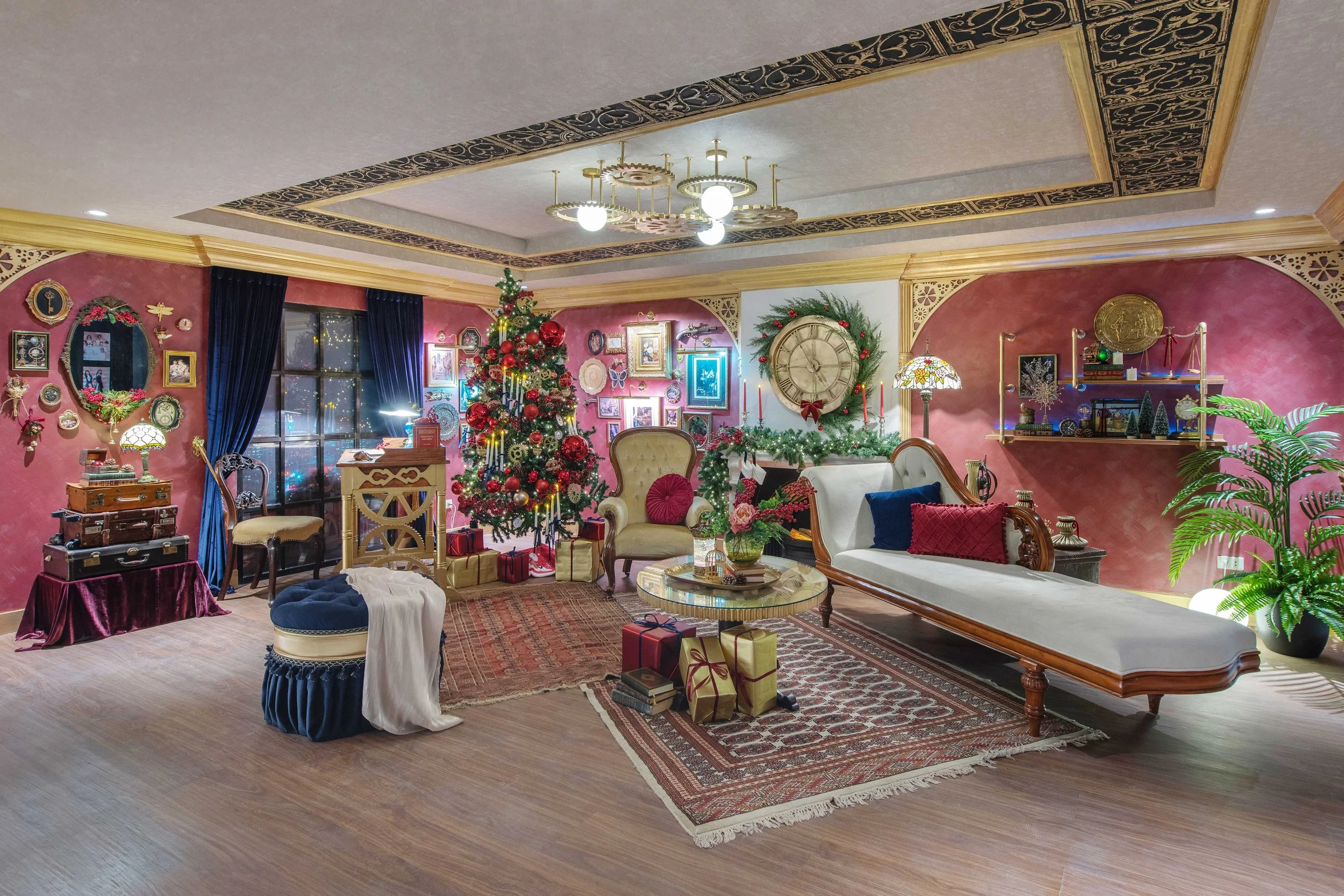 A cozy living room decorated for Christmas with a decorated Christmas tree, wrapped presents, and holiday decorations on the walls and shelves. The room has pink walls, a large clock with a wreath, and vintage-style furniture.