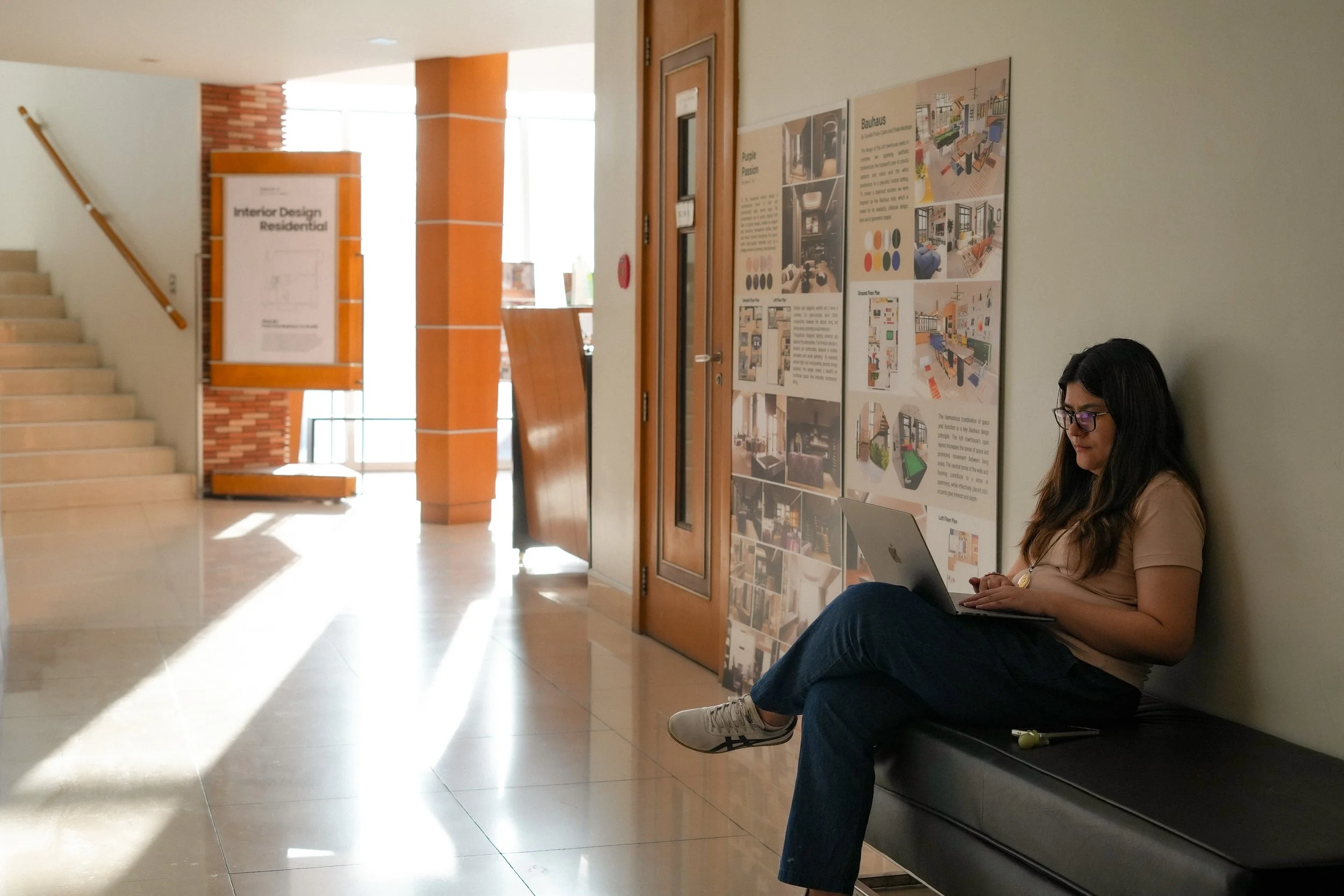 A woman wearing glasses, a beige t-shirt, and jeans is sitting on a bench with a laptop on her lap in a well-lit indoor public space. She is leaning against a wall near some posters or informational boards.