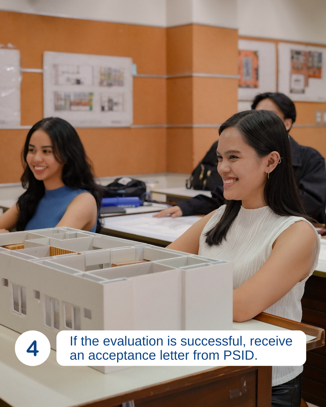 Students in a classroom sitting at desks, smiling, with a architectural model in the foreground. One female student is wearing a white sleeveless top, and another is wearing a blue sleeveless top.