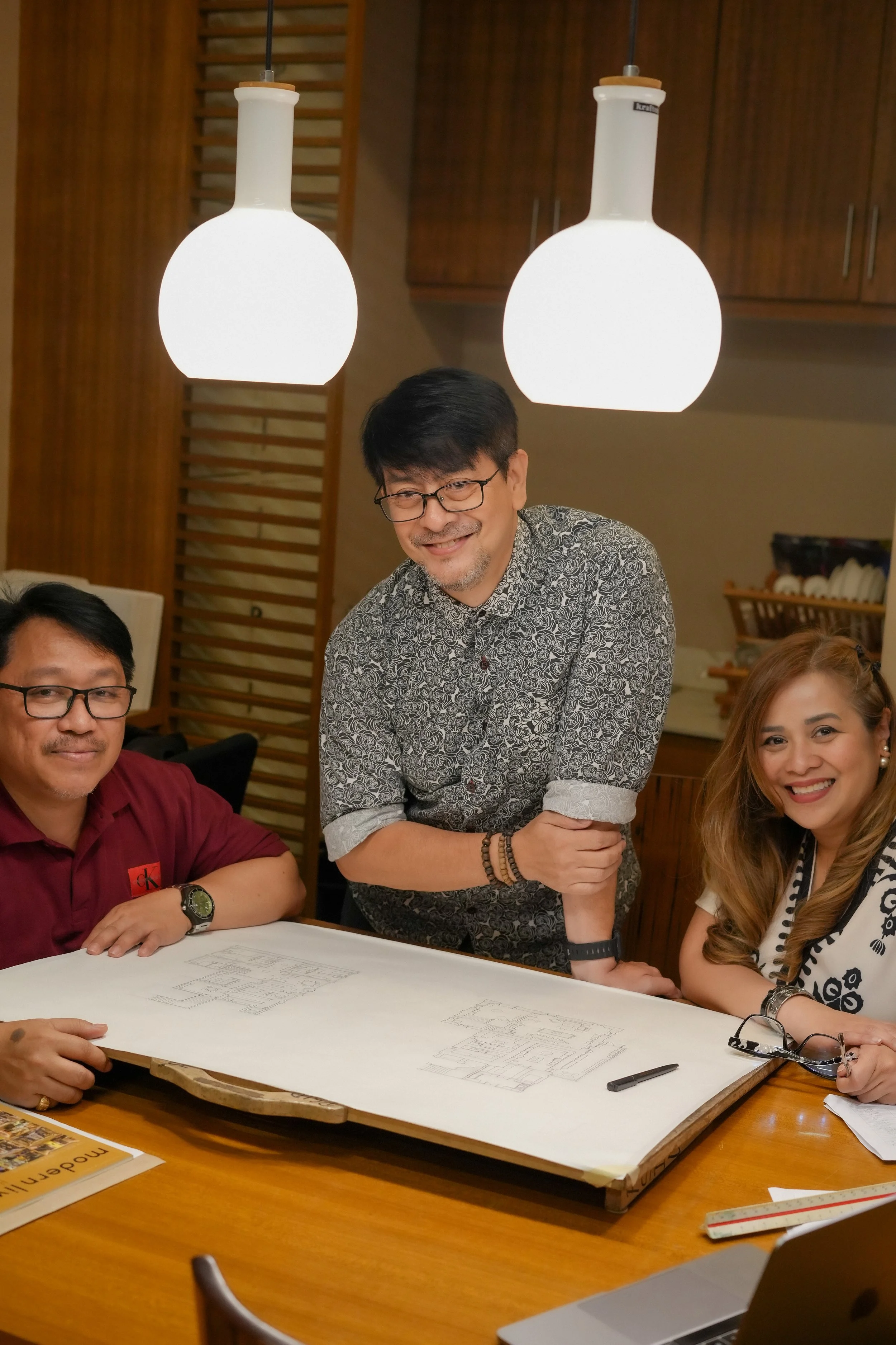 Four people gathered around a table with architectural drawings, with two hanging ceiling lights above, and a man in the center arm wrestling a woman on the right.