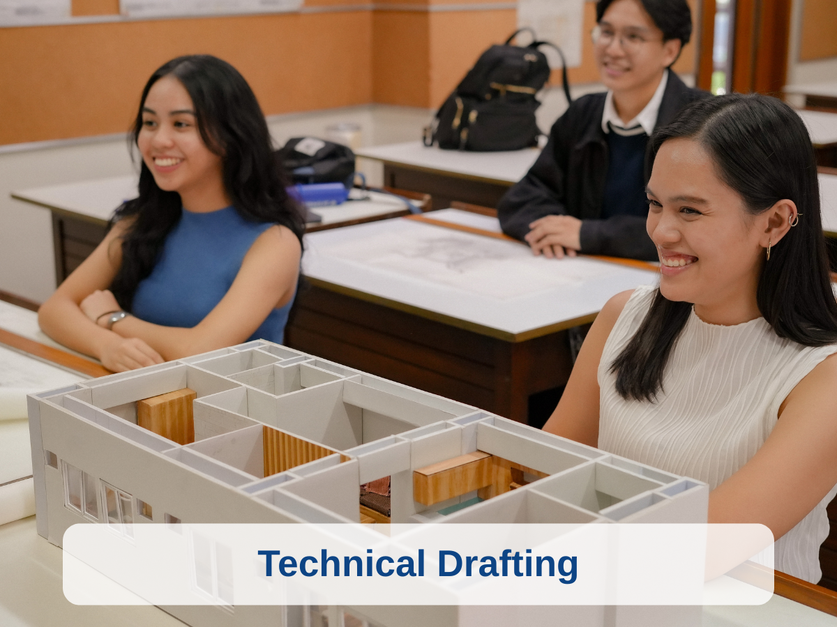 Three students in a classroom, sitting at desks with a model of a building's floor plan in front of them, engaged in technical drafting activities.