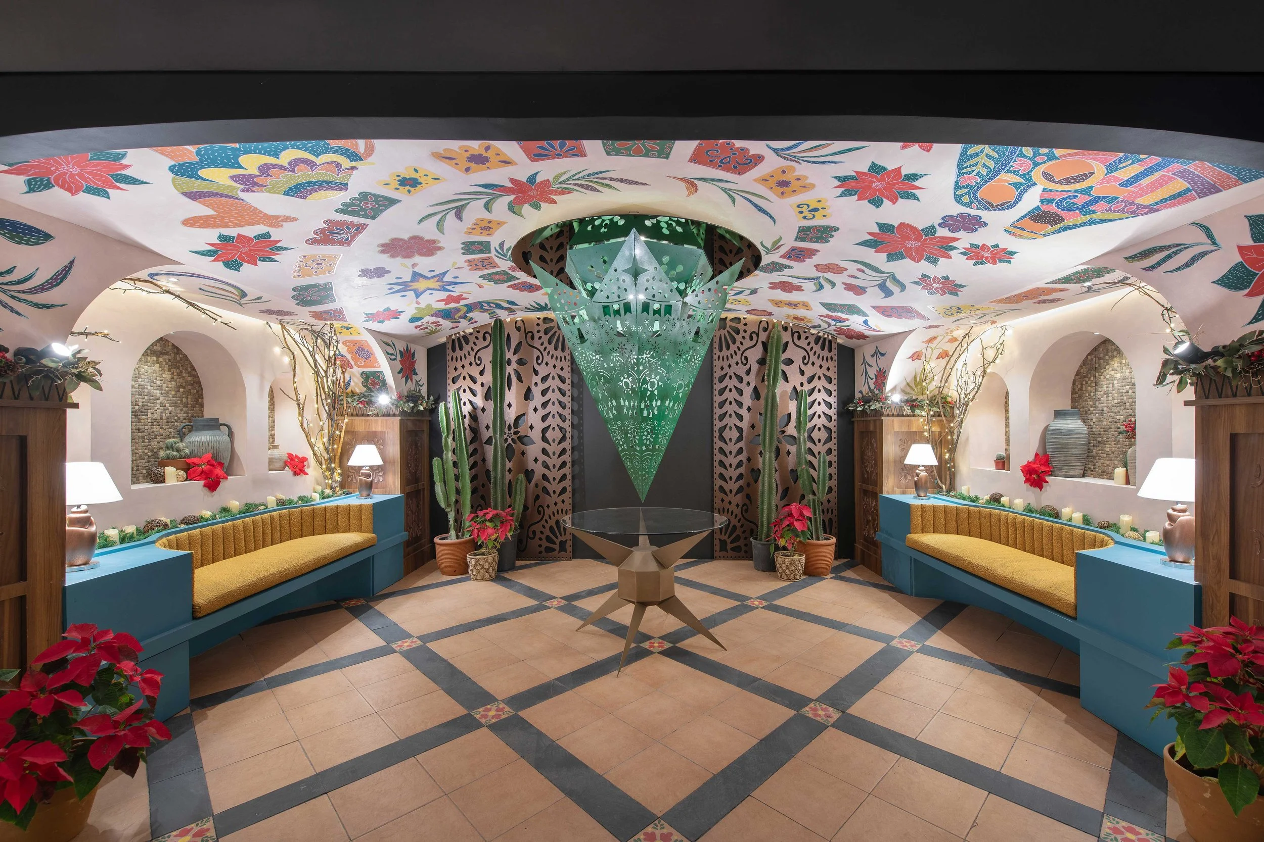 Indoor space with decorated ceiling featuring colorful floral and geometric patterns, round arches, plants, and seating areas with yellow cushions and blue benches.