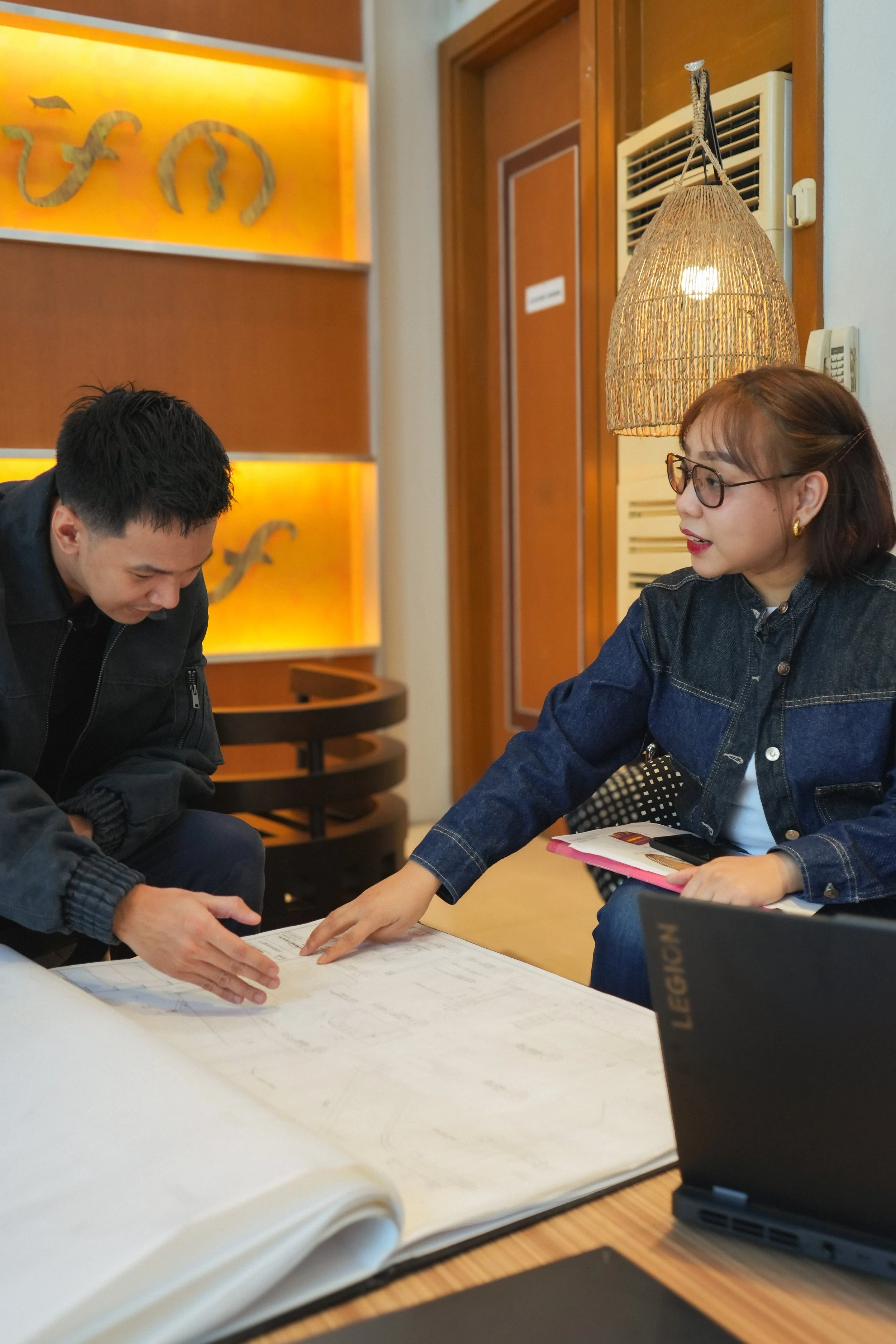 Two people are seated at a table, looking at documents. The woman on the right has short brown hair, wears glasses, a denim jacket, and is pointing at the papers. The man on the left has short dark hair, wears a black jacket, and is leaning forward. There are papers, a laptop, and a pink notebook on the table. Behind them is a decorative wall with orange lighting and snake artwork, a door, and a wall-mounted air conditioner. A wicker lampshade hangs above them.