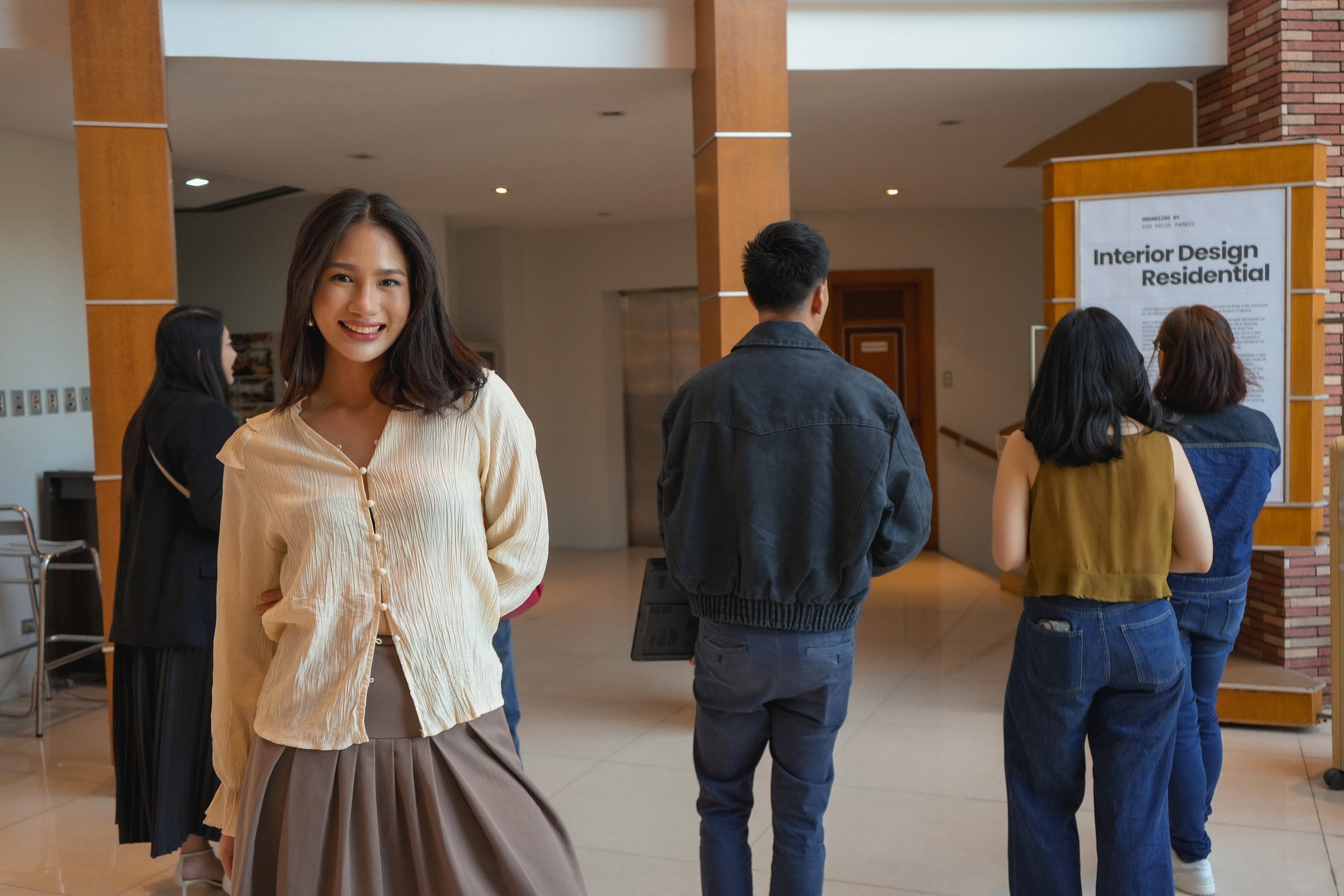 A group of people standing indoors, with a young woman smiling at the camera in the foreground. A sign on the wall reads 'Interior Design Residential.'