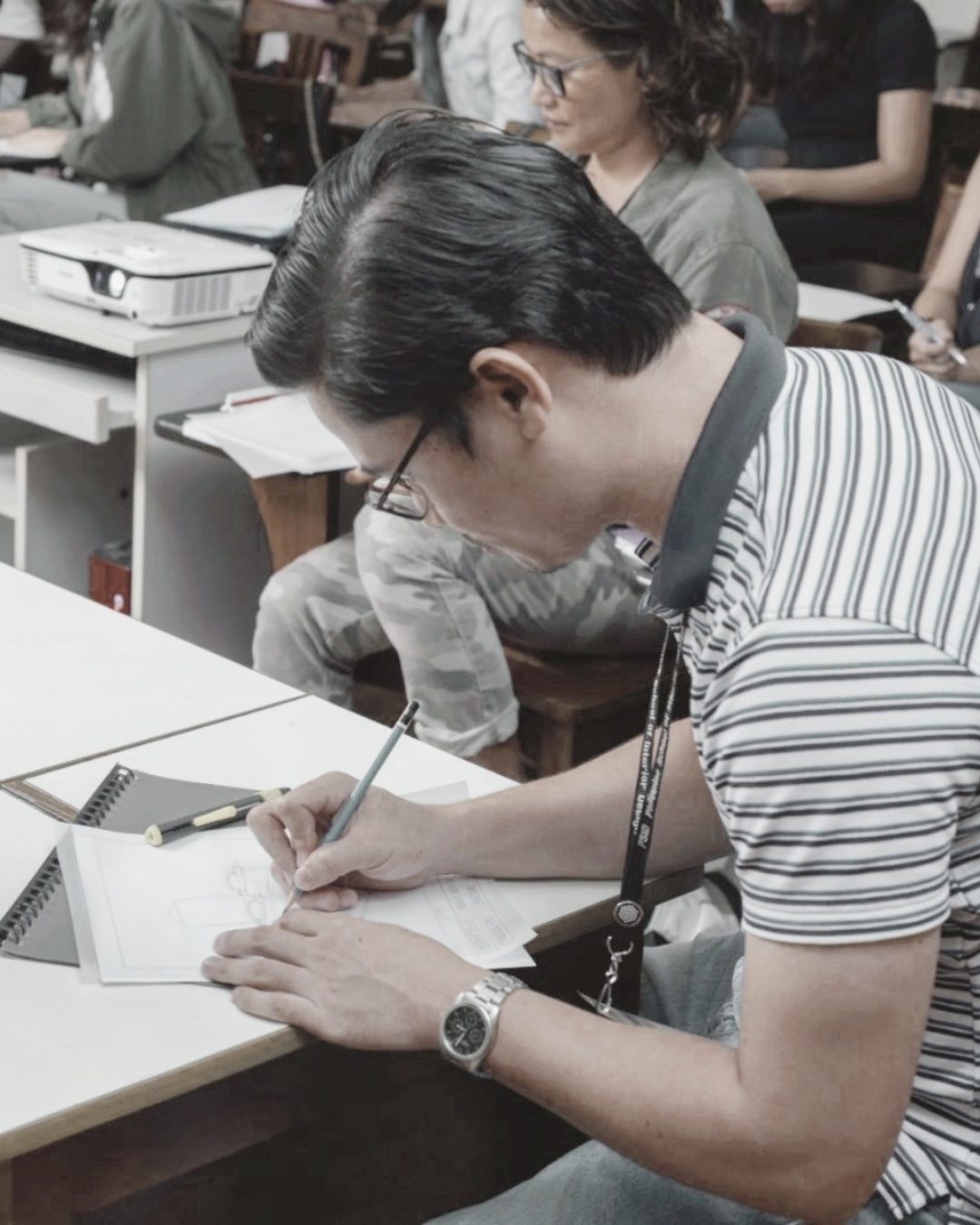 A man sitting at a desk, drawing on paper with a pencil, wearing glasses, a striped shirt, and a watch, in a classroom or workshop setting with other people and a projector in the background.
