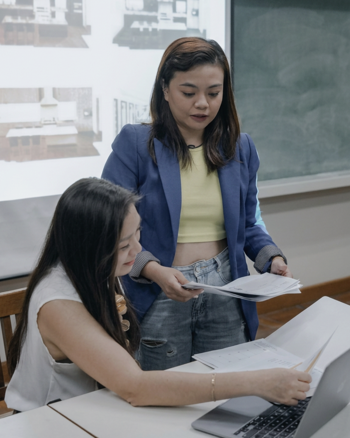 Two women in a classroom or office, one sitting at a desk using a laptop and the other standing, holding papers and looking at the laptop screen.