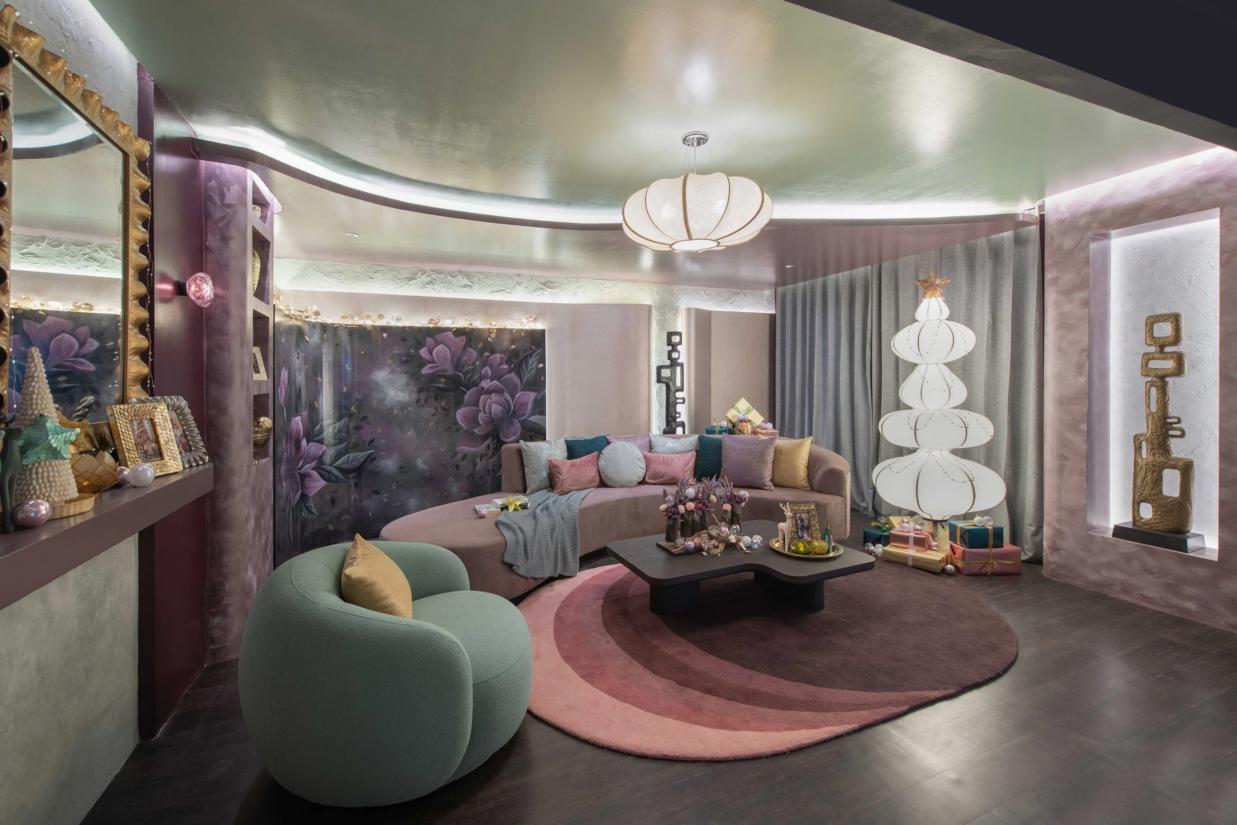 A cozy living room decorated with pastel colors and soft lighting. There is a large pink sectional sofa with various cushions, a dark coffee table with small decorated gifts, and a pink and purple floral painting on the wall. The room has unique lighting fixtures, including two modern pendant lamps and decorative lights along the ceiling edges. The space is accented with gold and glittery decor, and presents are wrapped on the floor.
