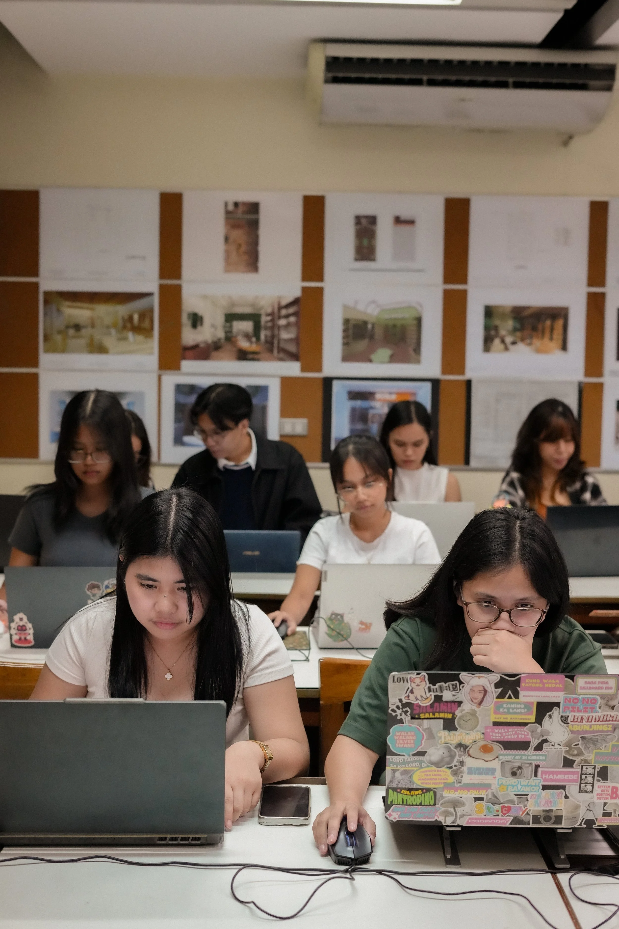A group of young women working on laptops in a classroom or computer lab, with a bulletin board displaying photographs or artwork in the background.