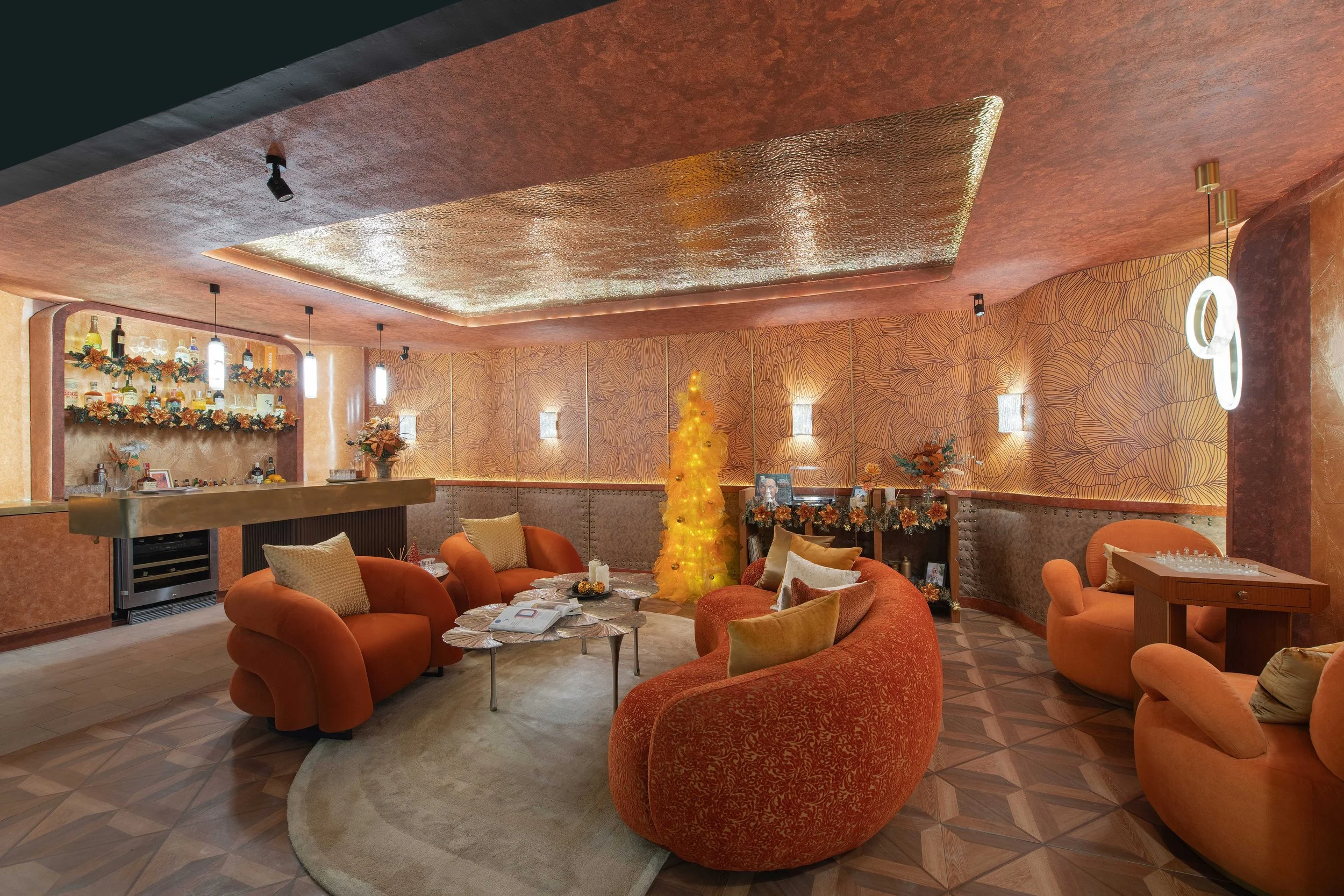 A cozy, decorated lounge area with orange and red velvet furniture, a small white circular table, a Christmas tree with gold decorations, and a bar area with bottles and holiday garlands, illuminated by modern hanging lights.