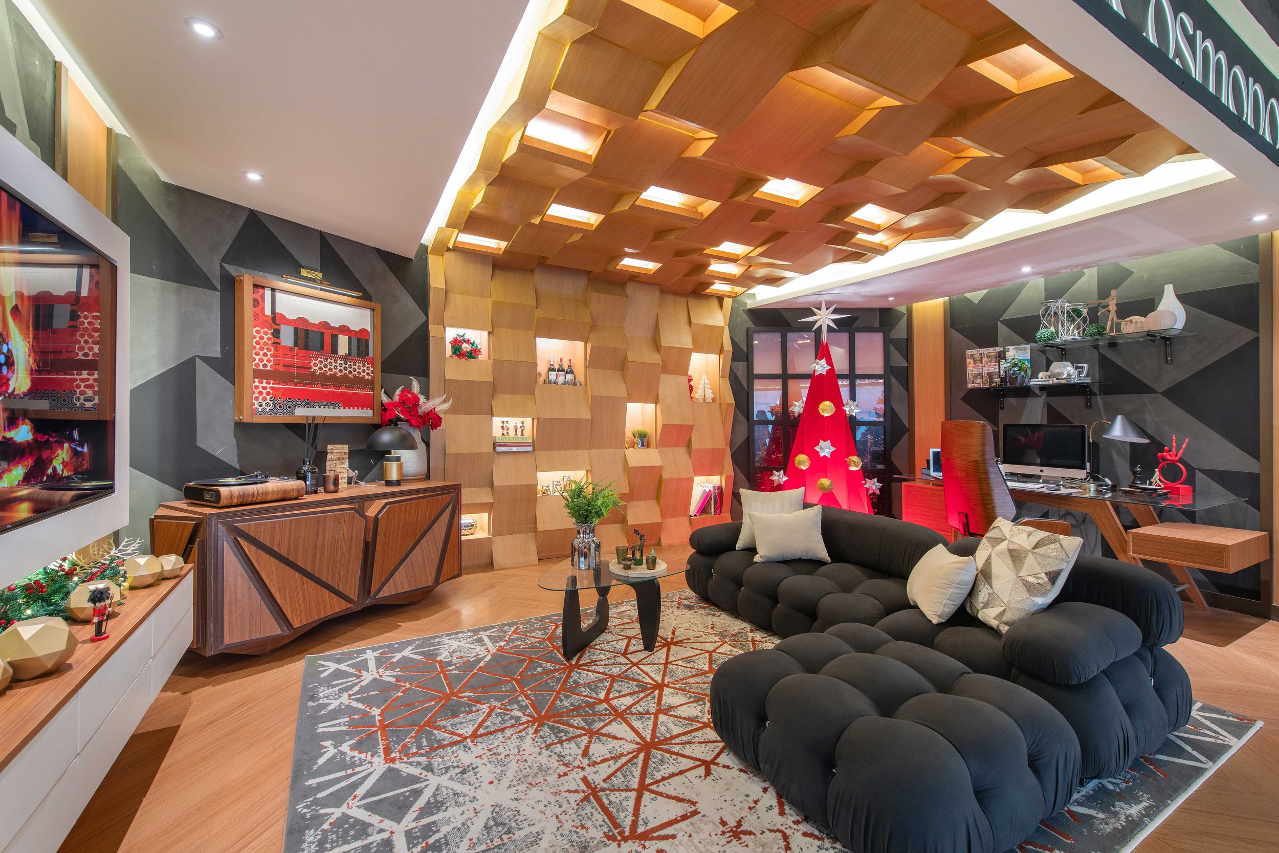 A modern living room decorated for Christmas with a small artificial Christmas tree with star ornaments and gold decorations, a black tufted sectional sofa with throw pillows, a geometric patterned rug, a wooden coffee table, wall-mounted shelves with decorative items, and a ceiling with a wooden architectural design with built-in lighting.