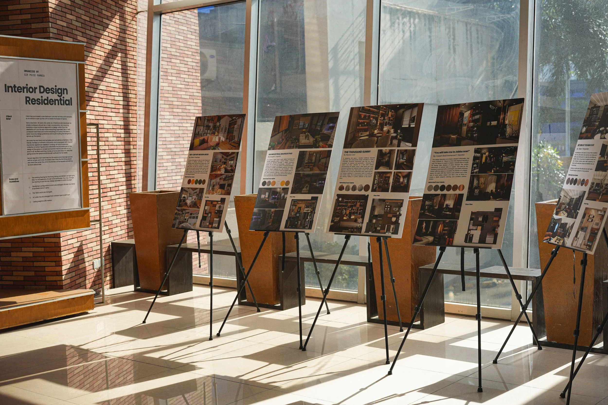 Exhibition display boards featuring interior design projects indoors near large windows with brick and concrete walls, sunlight casting shadows on the tiled floor.