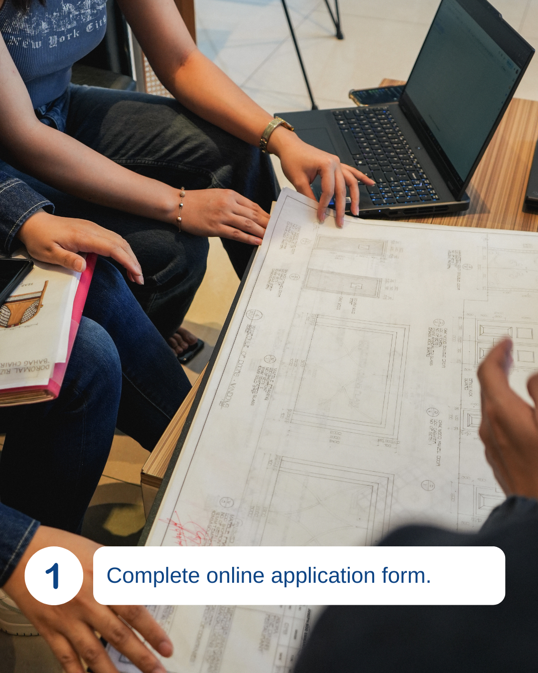 People reviewing blueprints and filling out application forms at a desk with a laptop.