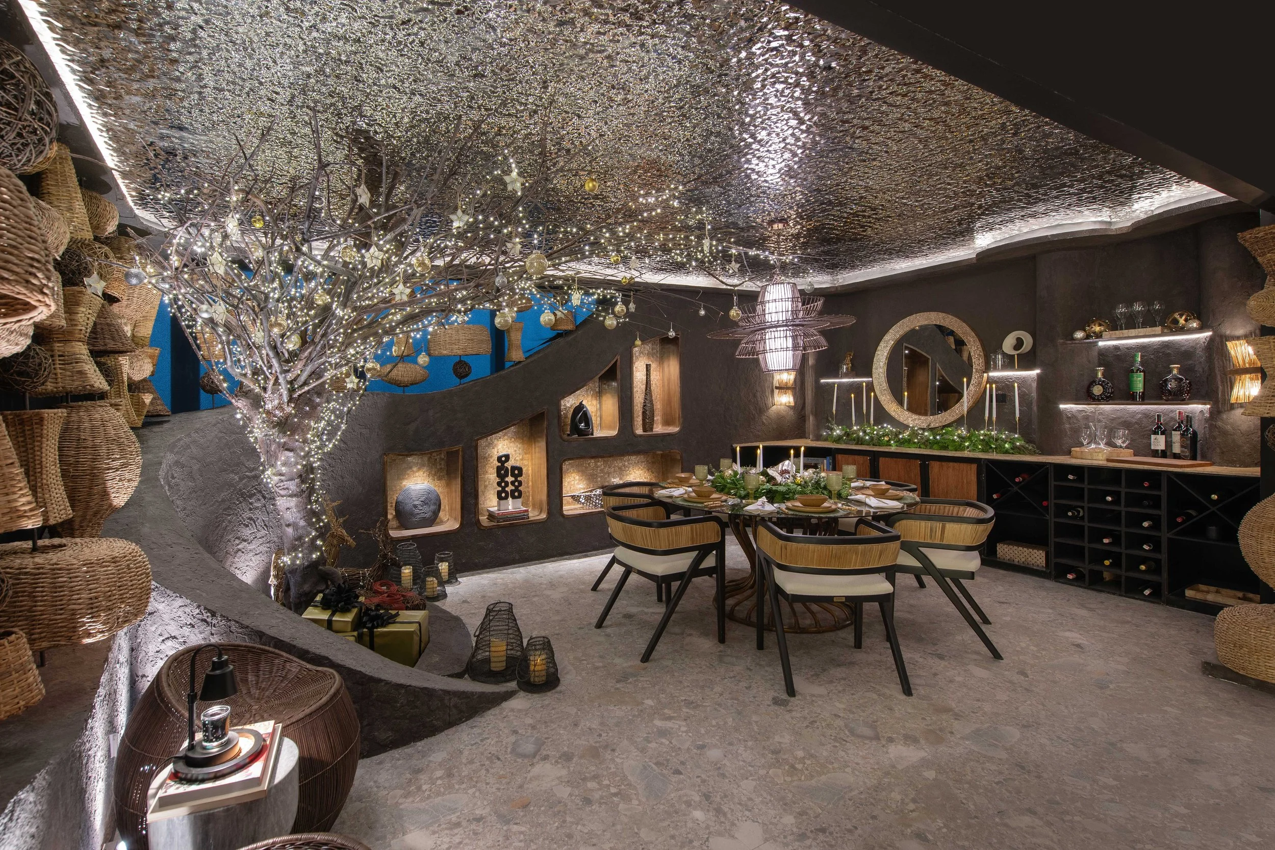 A decorated indoor dining space with a tree wrapped in fairy lights and ornaments, surrounded by wicker baskets, shelves with art decor, a round dining table with chairs, and various lighting fixtures on dark textured walls.