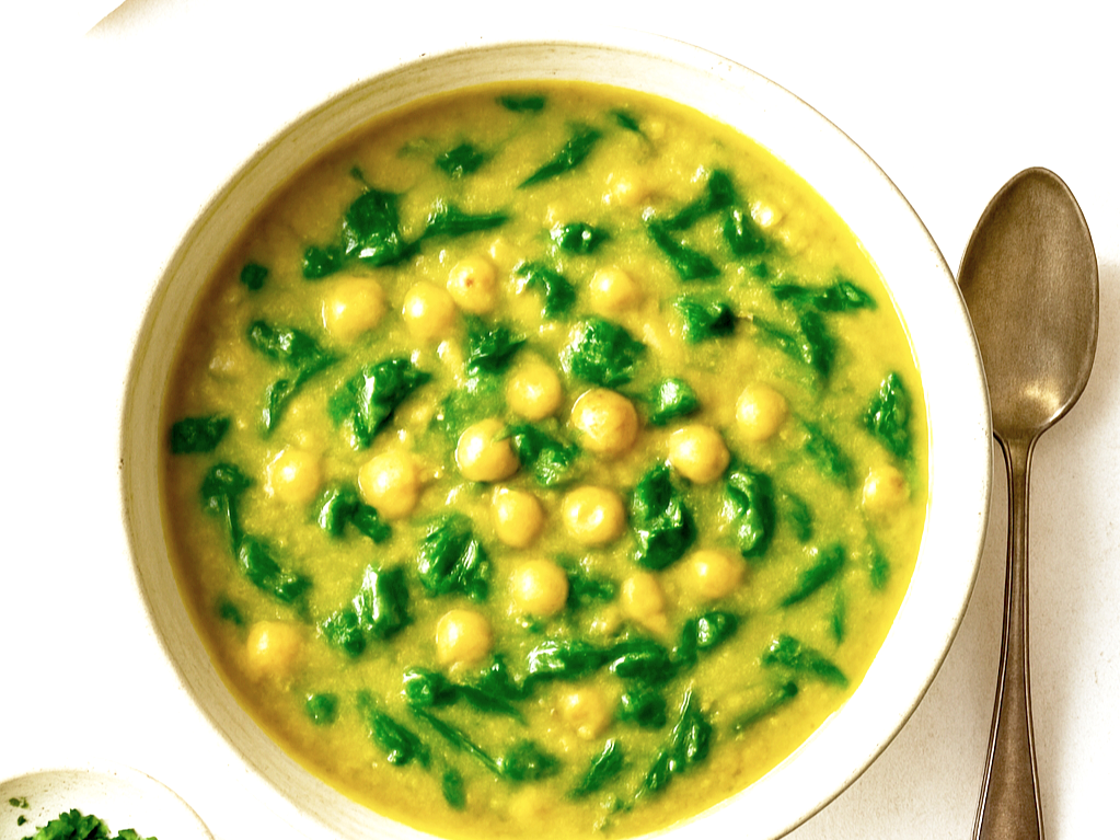 An image of chickpea & spinach soup in a bowl on a table with a spoon next to it.