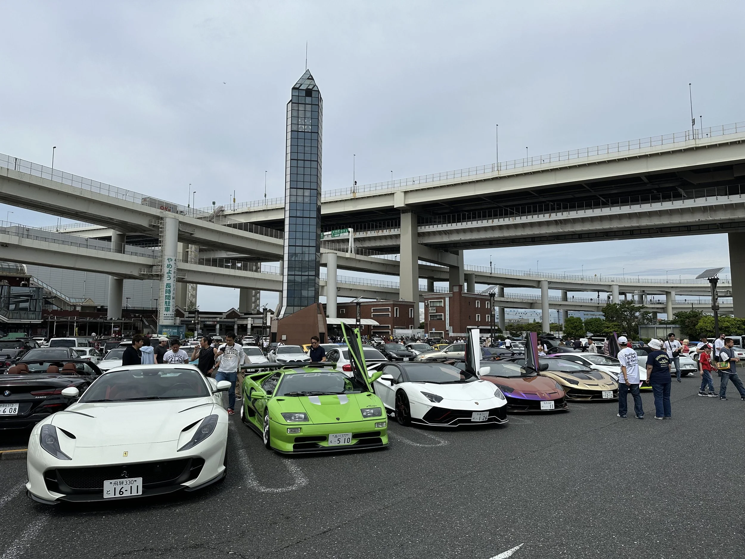 Exposition de voitures de luxe, incluant une Ferrari blanche, une Lamborghini verte, une Lamborghini blanche, une Lamborghini violette, et une Lamborghini or, sous un ciel nuageux, avec un grand nombre de personnes autour dans un parking urbain.