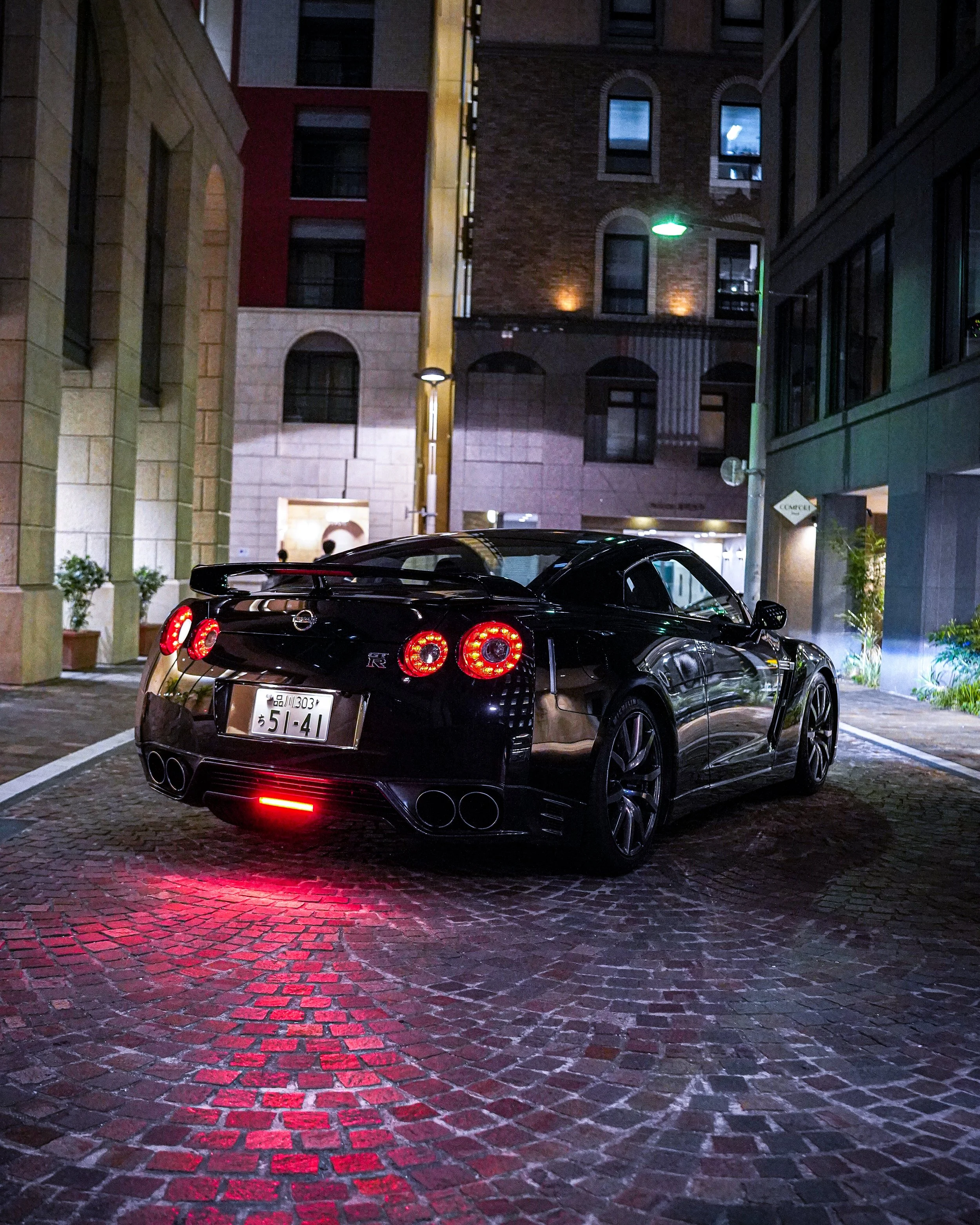 A black Nissan GT-R sports car parked in a cobblestone alley at night, illuminated by streetlights and building lights.
