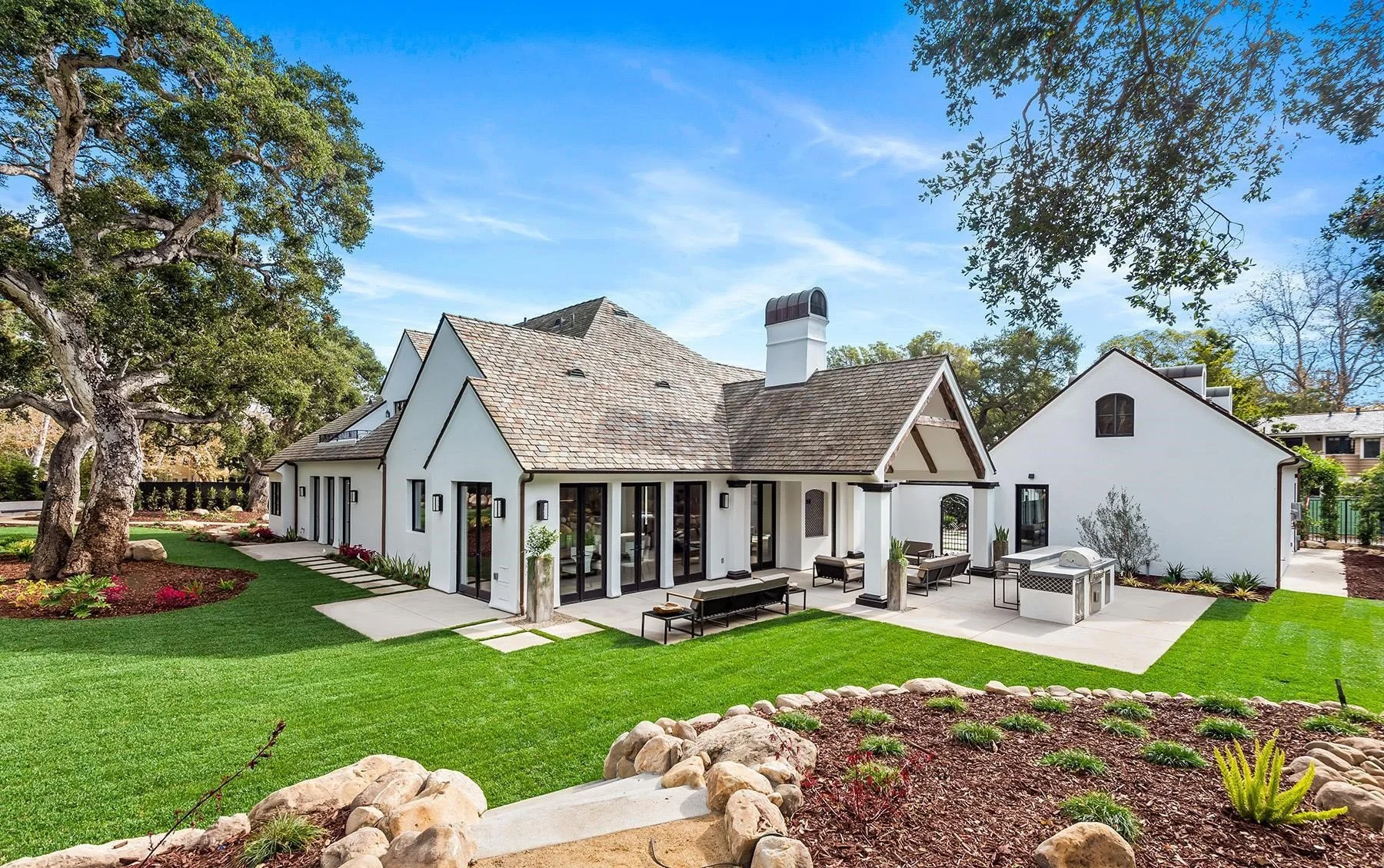 Modern white house with black trim, large glass doors, outdoor seating area, grill, lush green lawn, trees, and landscaped garden.