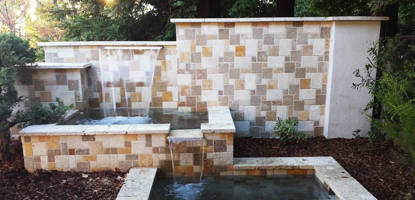 Stone water feature with small waterfall, rectangular pools, surrounded by mulch and greenery.