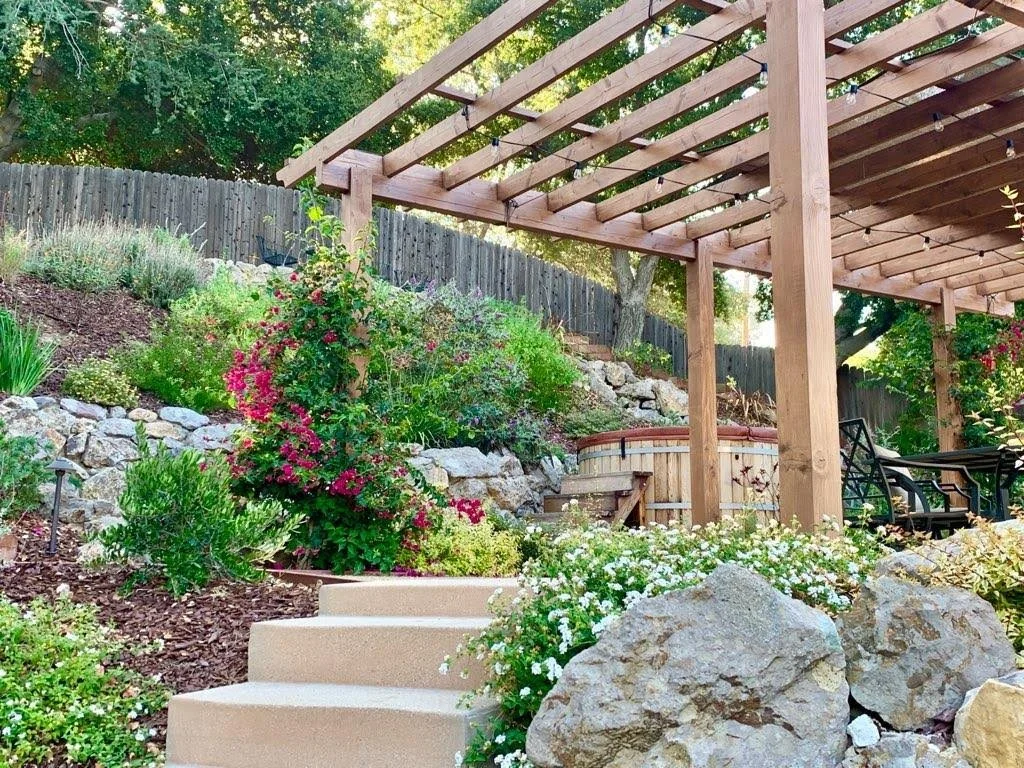 A backyard garden with a pergola, stone steps, colorful flowering plants, large rocks, and a wooden fence in the background.