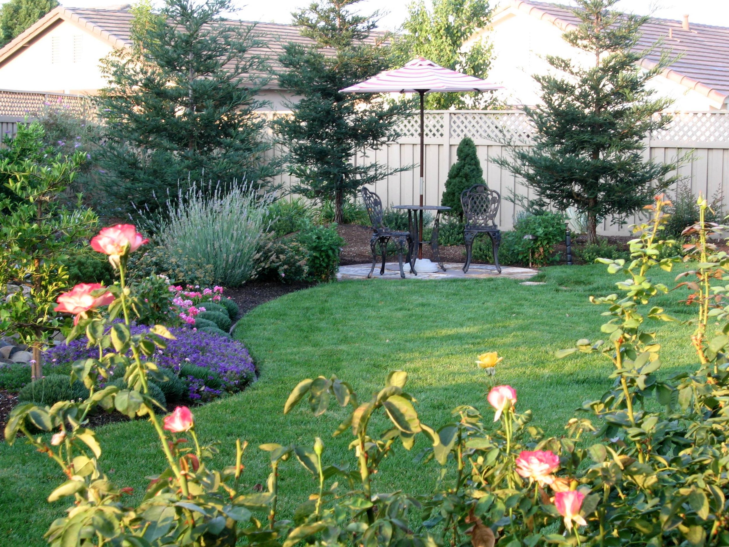 A backyard garden with a green lawn, colorful flowers, and three trees behind a fenced area. There is a small patio with a table, two chairs, and an umbrella.
