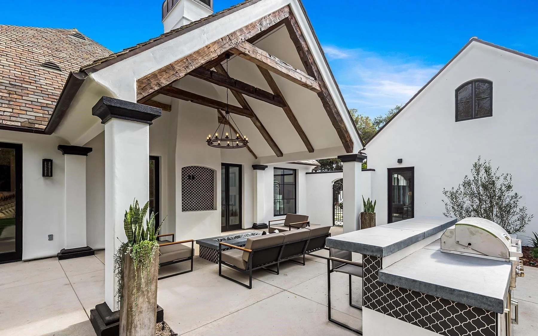 Modern outdoor patio with seating, potted plants, and built-in grill area under a wooden gable roof.