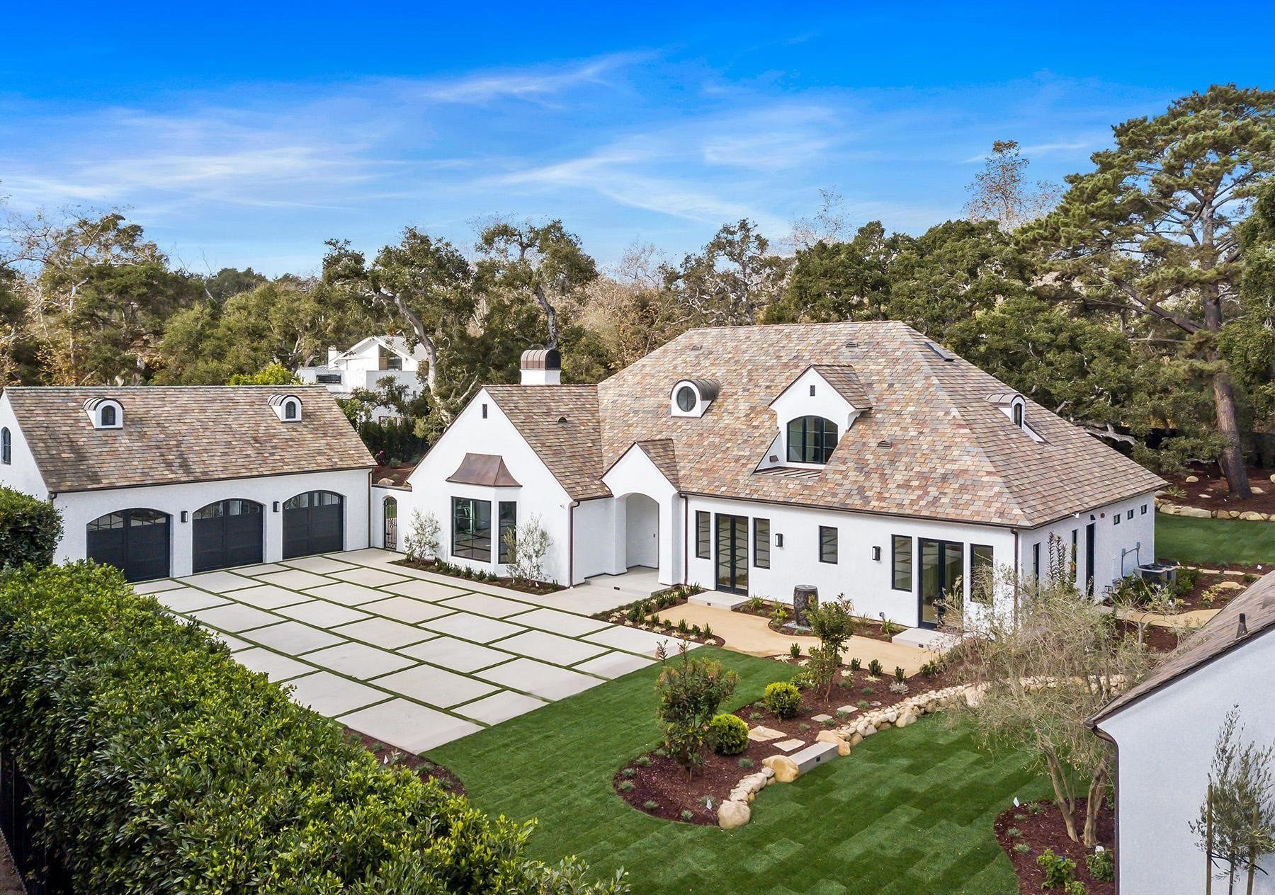 A large white house with a multi-gable roof, multiple windows, and a three-car garage. The front yard has a pattern of concrete slabs surrounded by grass, with small trees and landscaped garden beds.