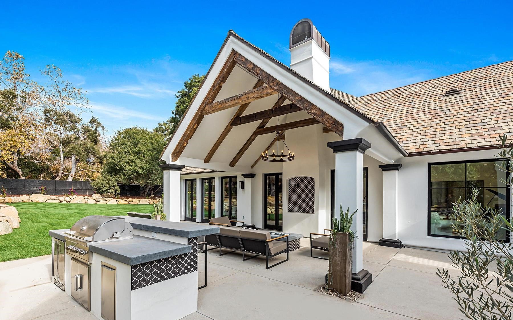 Modern outdoor patio with built-in grill, outdoor seating, and a chandelier under a pitched roof with exposed wooden beams at a well-maintained backyard.