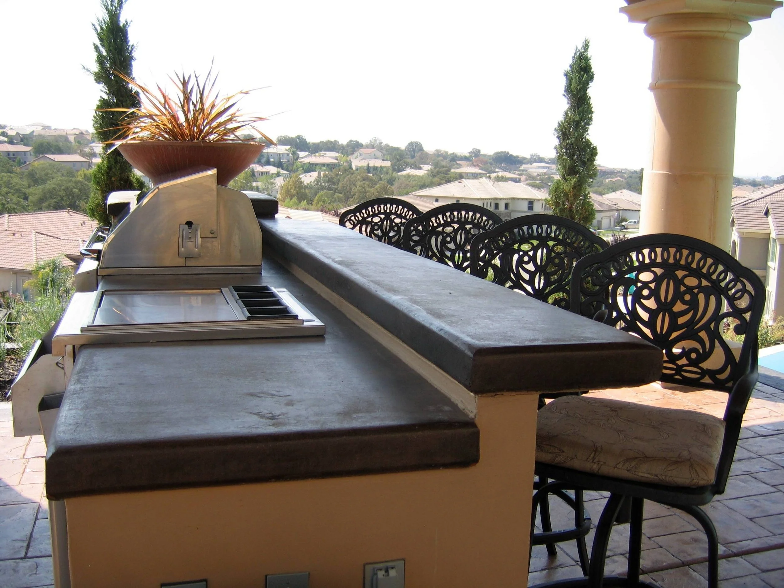 Outdoor kitchen with a built-in grill, counter space, and decorative chairs on a patio overlooking a neighborhood.