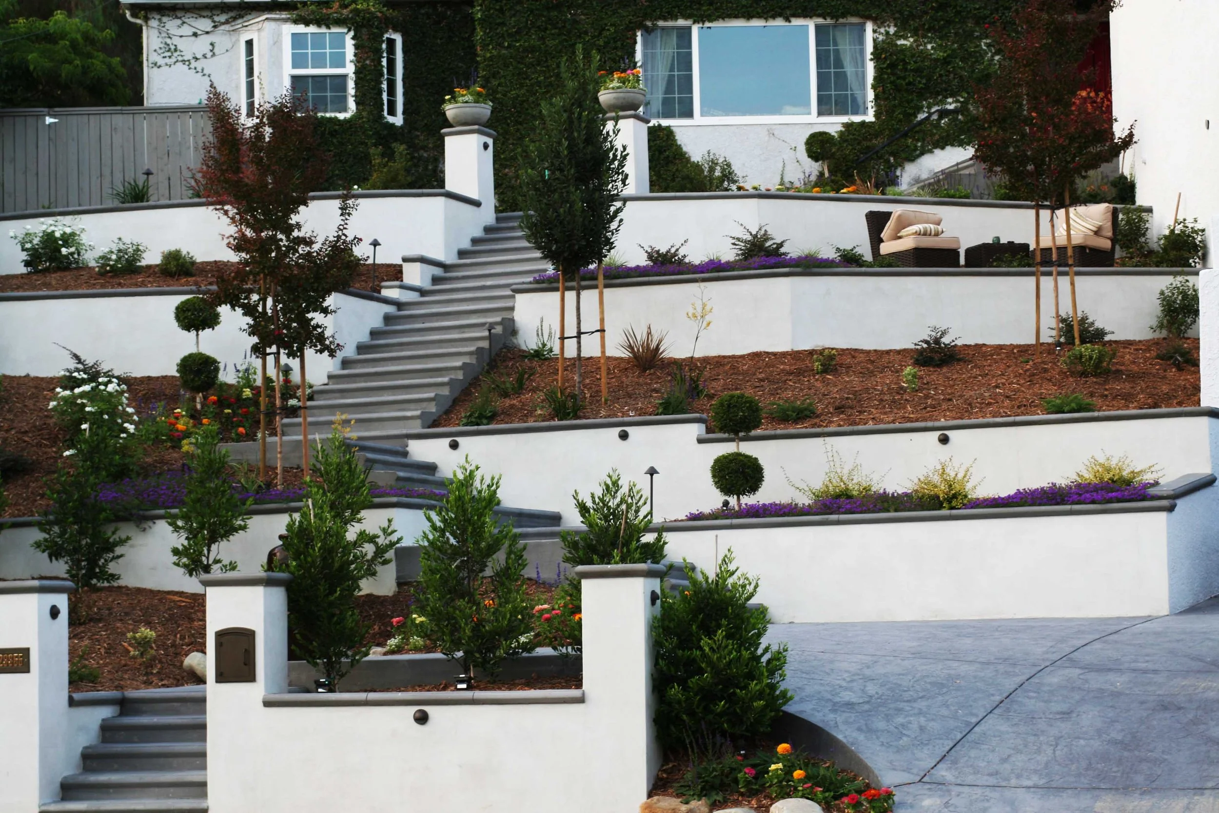 Multi-level white terraced garden with plants, small trees, and outdoor seating beneath a house with large windows.