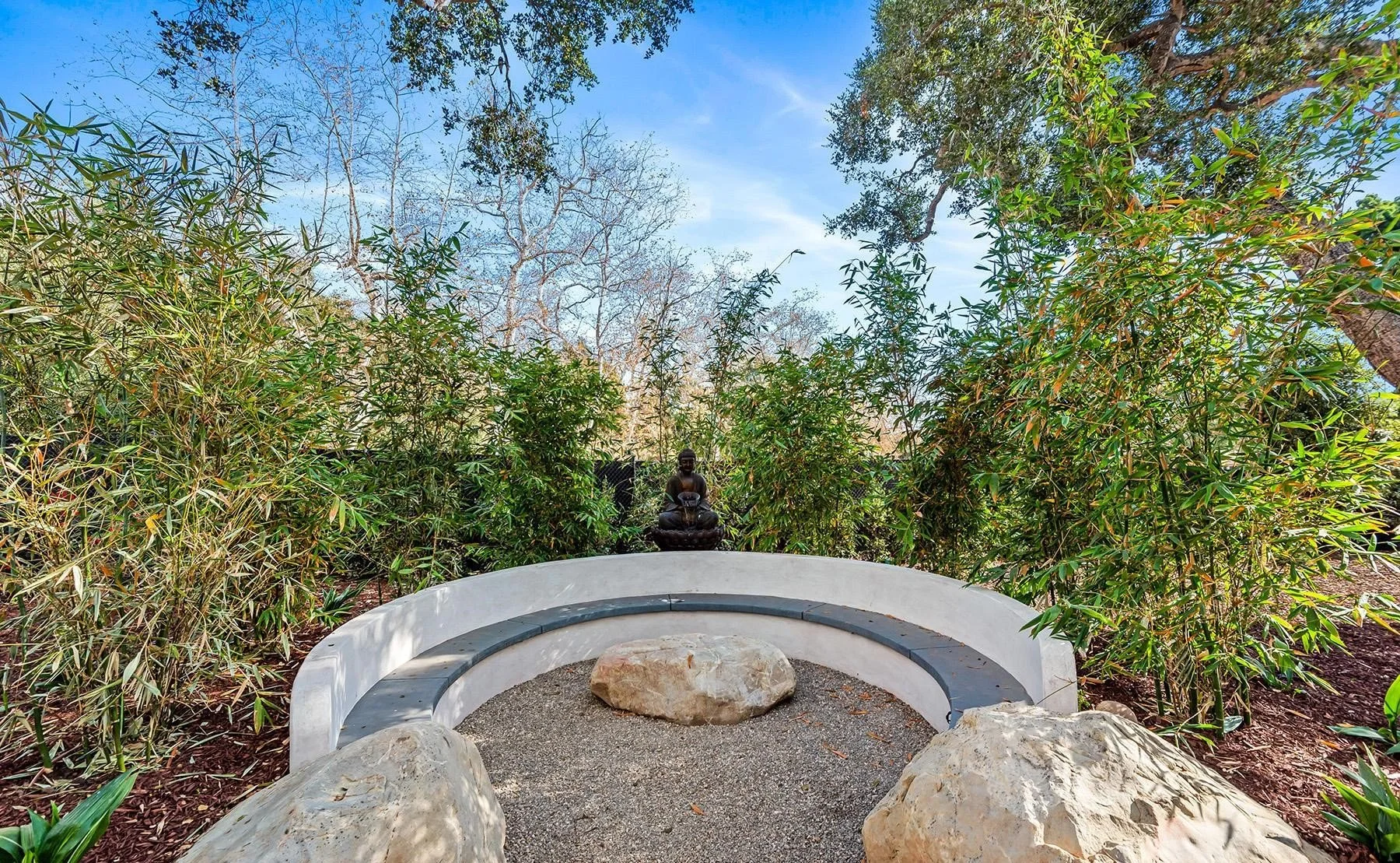 A small, circular outdoor seating area with a white wall and gray bench, surrounded by lush green plants and trees, with a small bronze statue of a seated person at the back and large rocks in the foreground, under a blue sky.