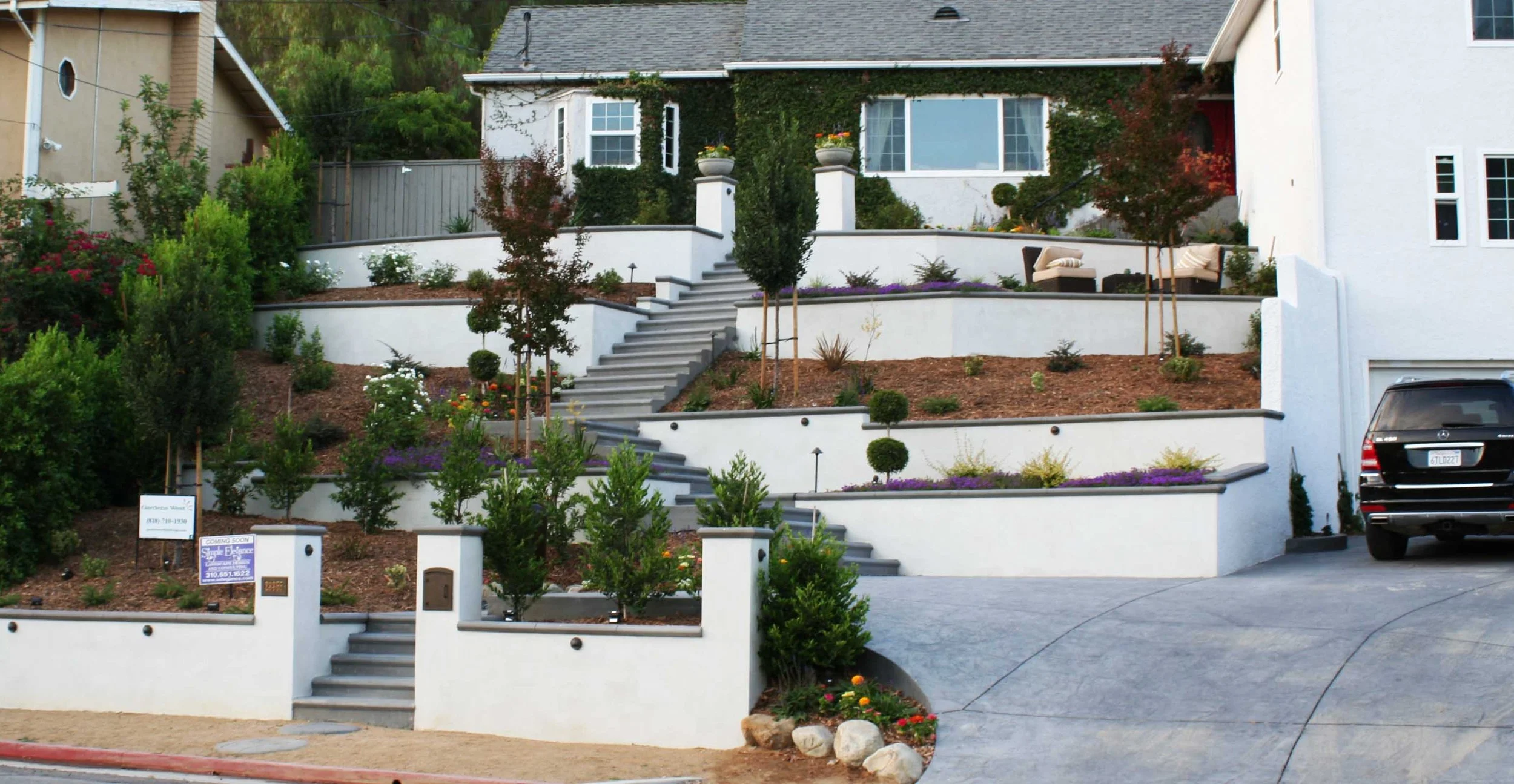 Multi-level white residential front yard with stairs, landscaped garden, trees, and seating area, including a driveway with a parked black vehicle.