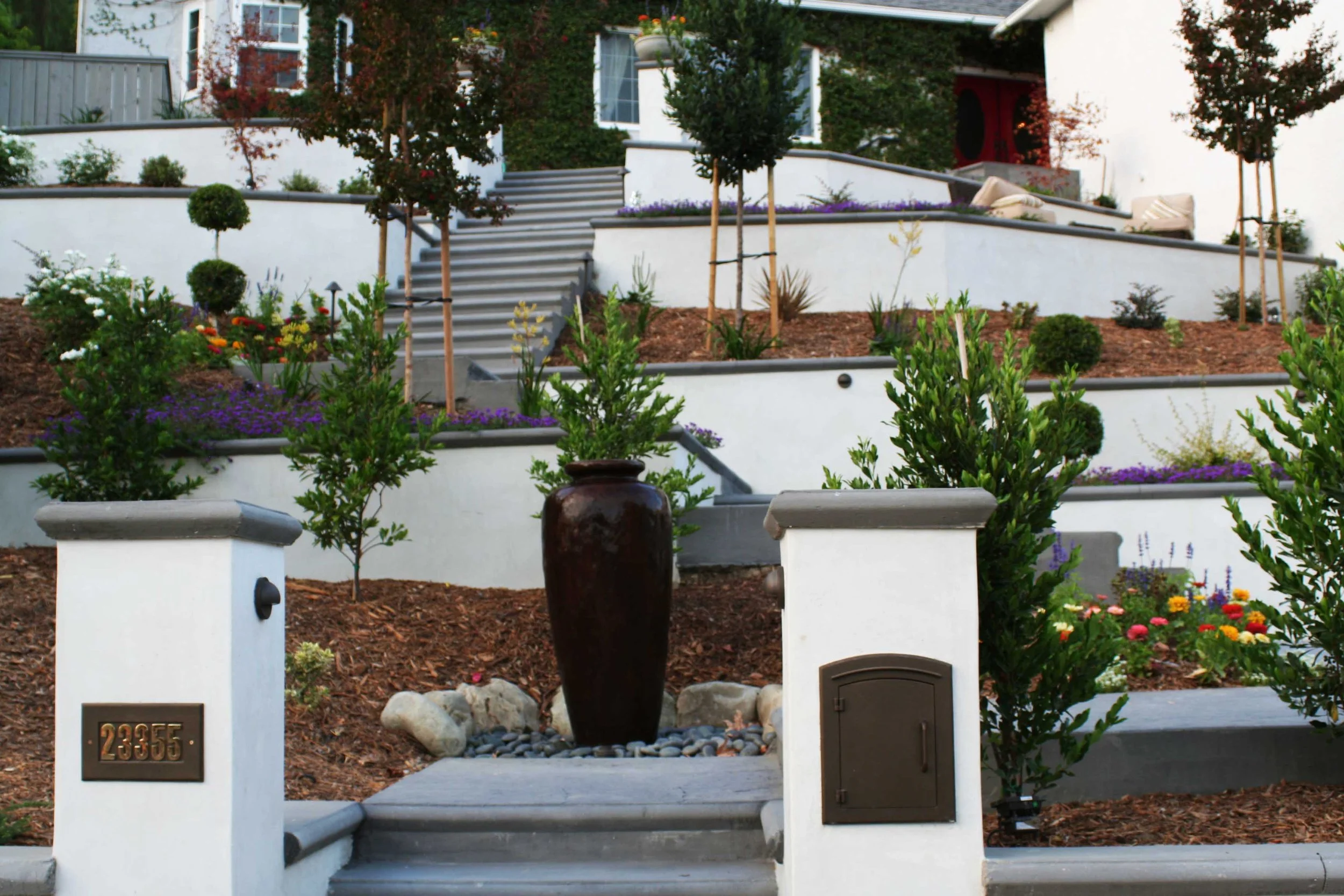 Multi-level landscaped front yard with white retaining walls, steps, and a variety of plants, flowers, and small trees, with a large dark vase centerpiece at the entrance.