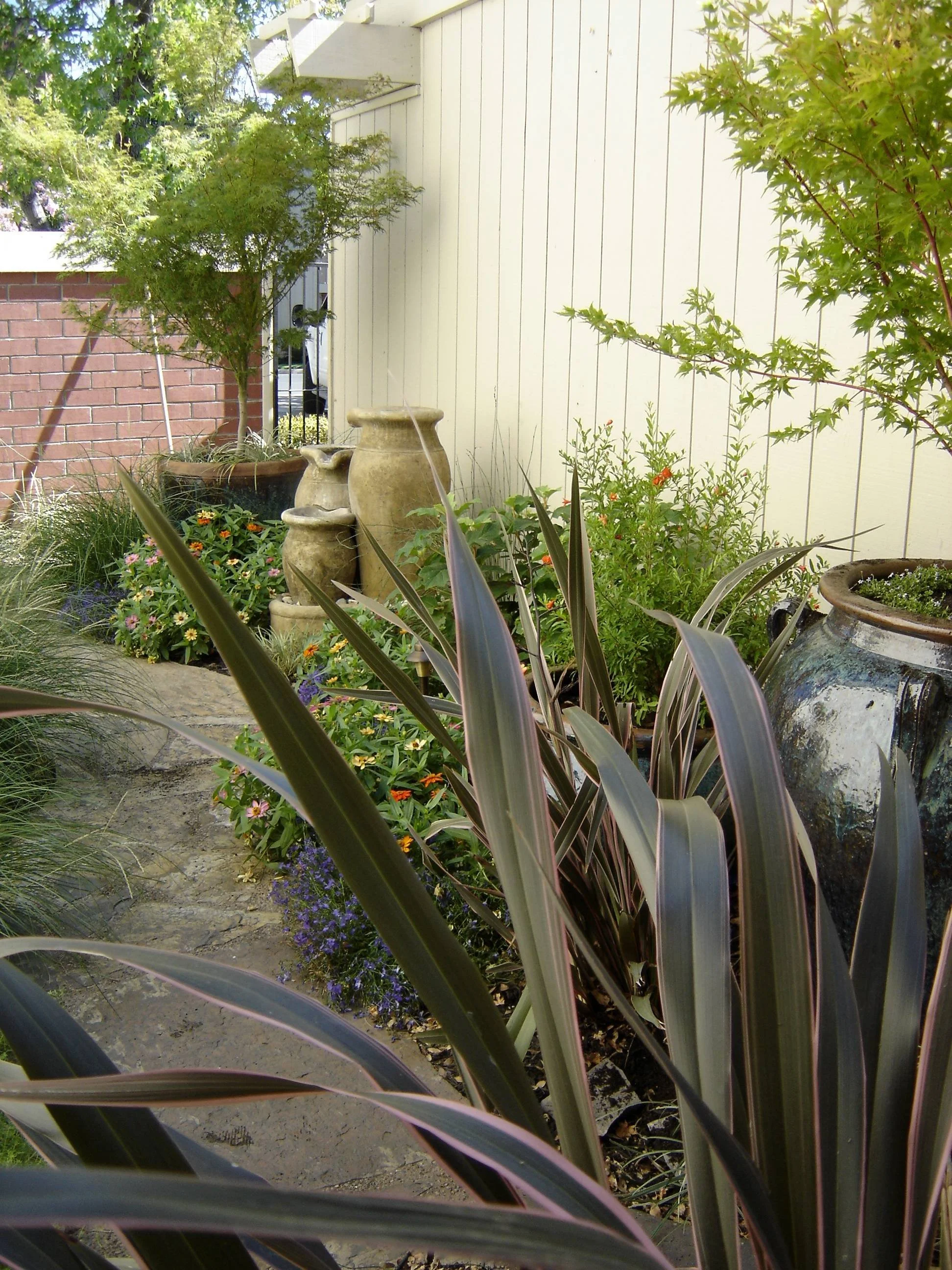 A garden with tall, purple, and green plants, decorative pots, and flowering shrubs beside a white wooden fence and house wall.