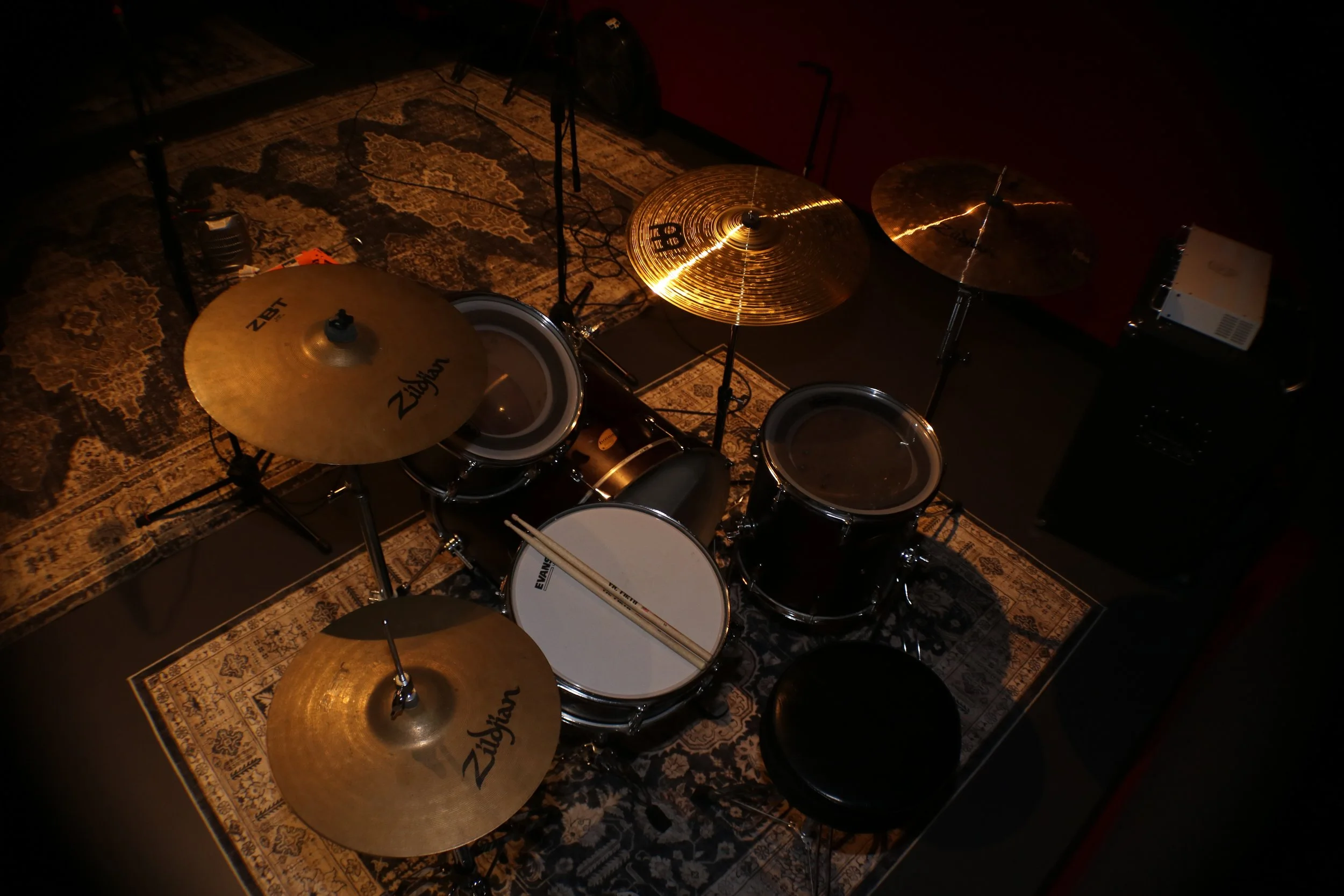 STUDIO 1 Music rehearsal studio with drum set, microphones, guitar, amplifiers, and rugs on the floor, dimly lit with ceiling lights and standing lamp.