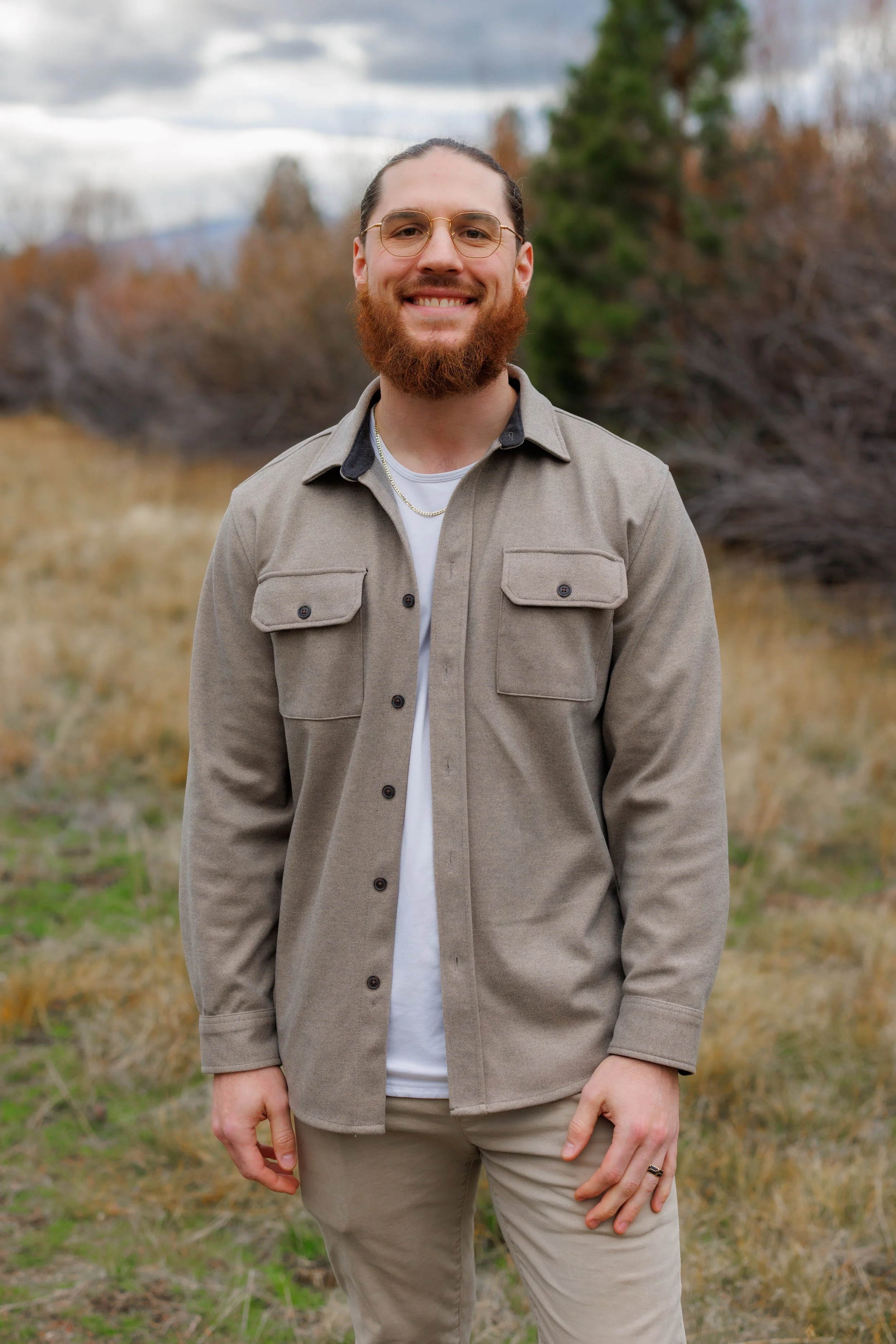 A young man with blonde hair styled back, dressed in a white blazer and shirt, standing against a pink background, with his hand on his chin, in a thoughtful pose.