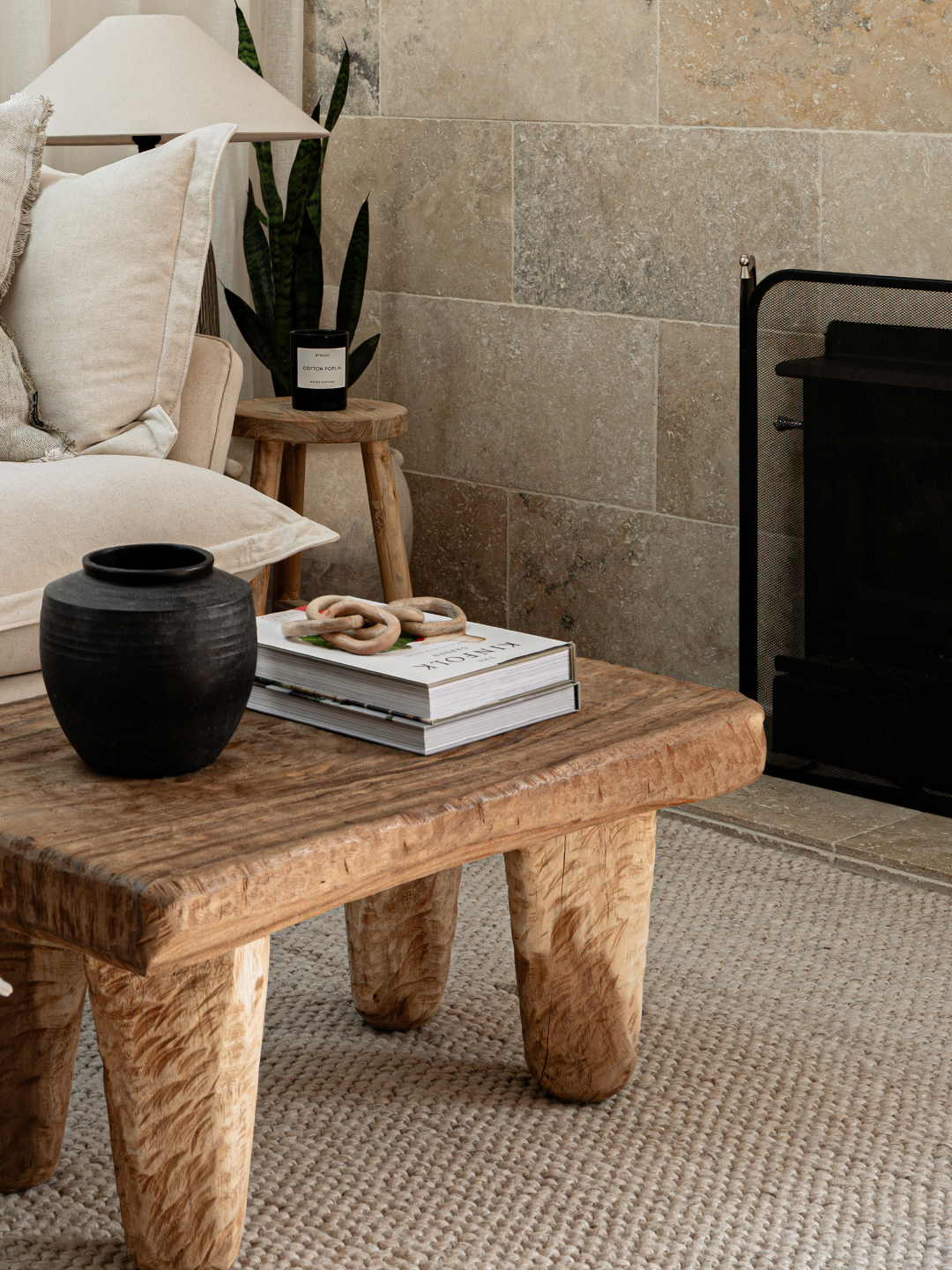 Sculptural timber coffee table styled with a black ceramic vase and books beside a travertine stone fireplace, Glenelg Retreat living room