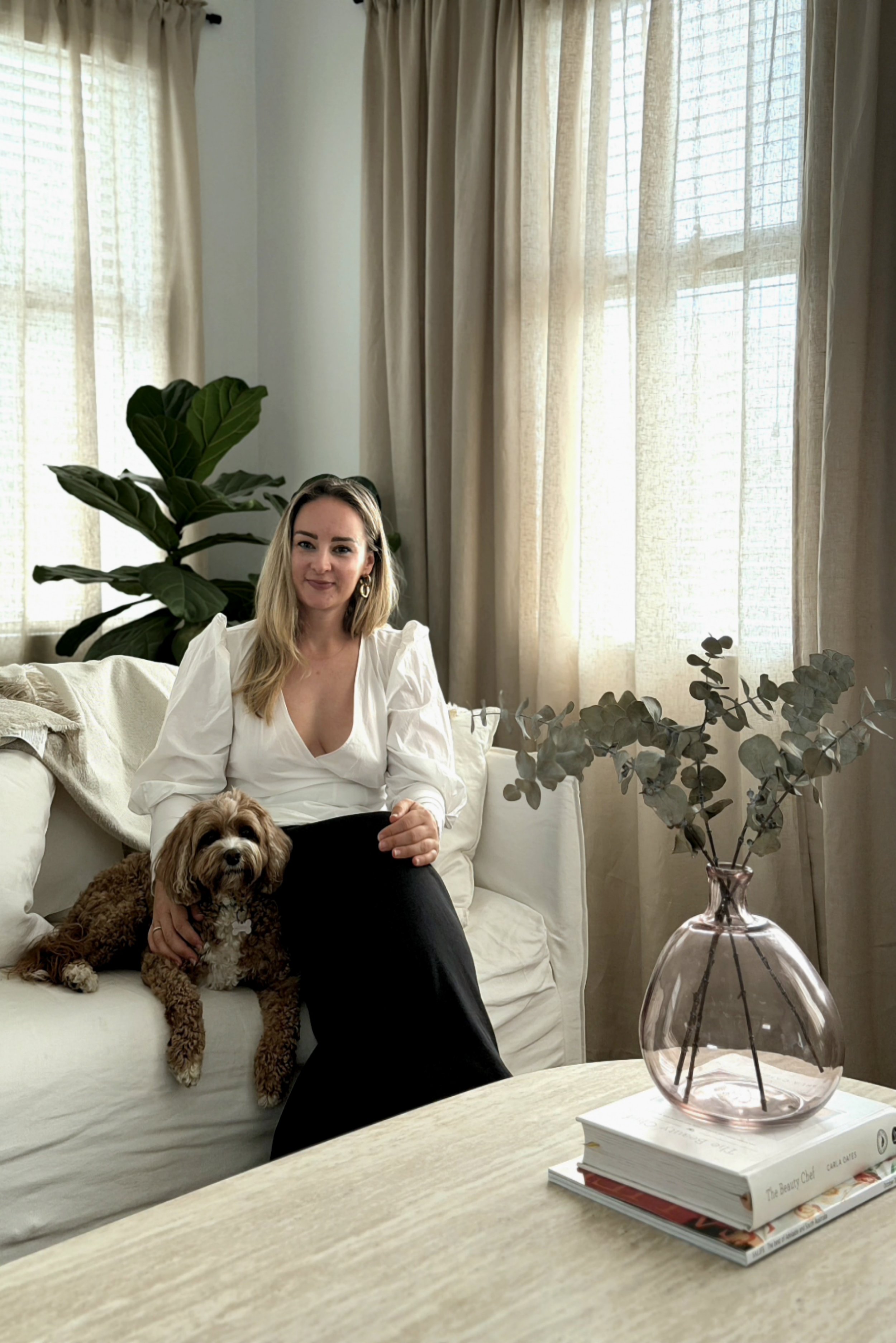 A woman wearing a white blouse and black pants sitting on a white couch with a small brown and white dog on her lap. The dog is lying down, and the woman is gently holding it. There is a large leafy plant in the background, and beige curtains covering a window. In the foreground, a round table holds a pinkish glass vase with dry eucalyptus stems, a white book, and a cookbook.