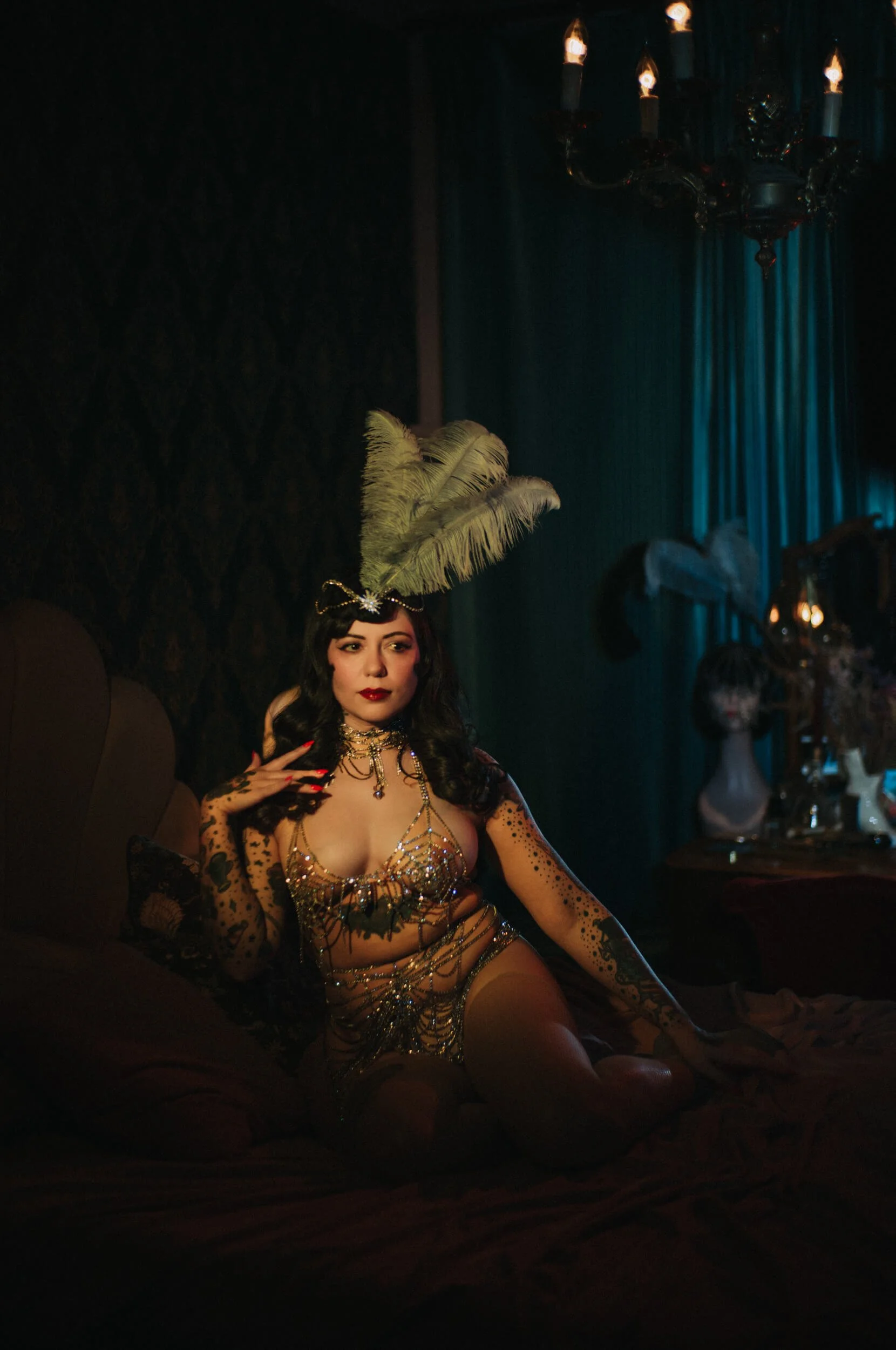 Bettie from Hell posing on a bed in a feathered headpiece, photographed with the Mandler 50mm f/1.4 E46 Pre-A in cinematic warm and teal lighting.