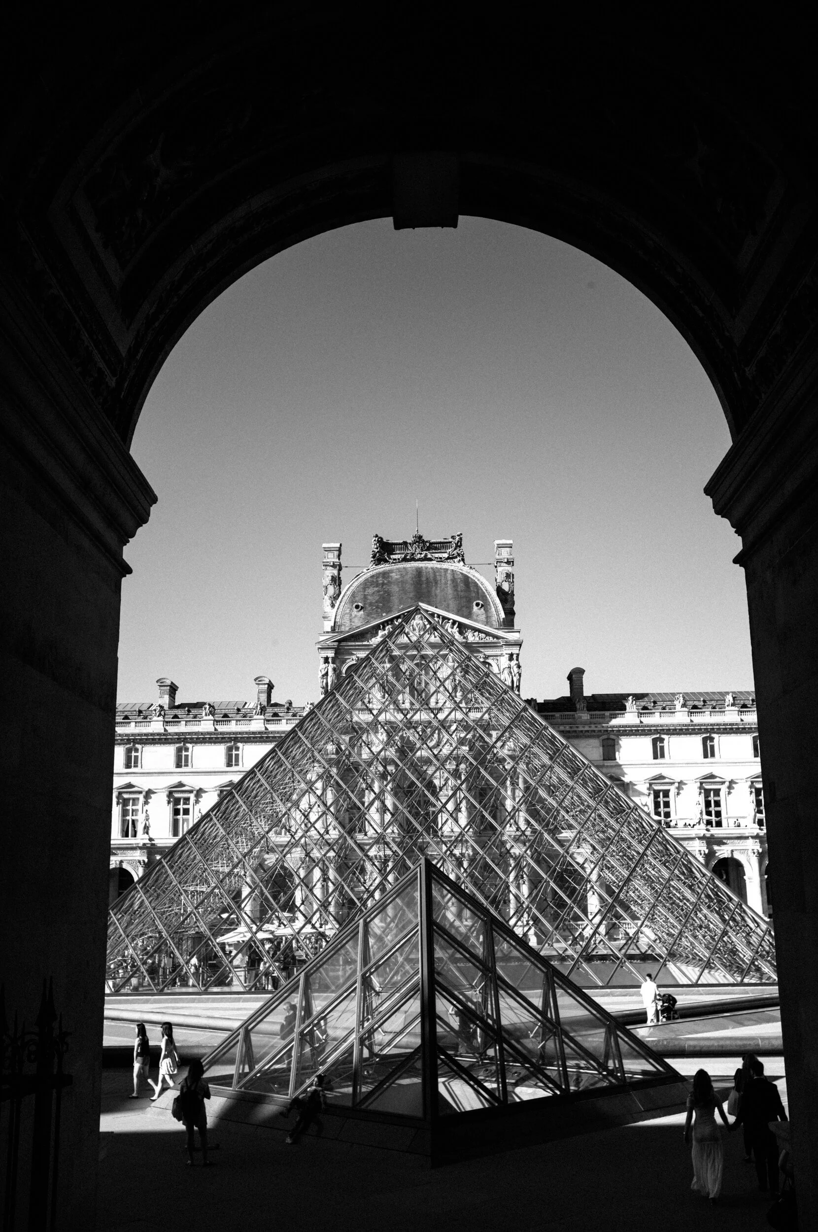 The glass Louvre Pyramid in Paris framed through a dark arch, photographed in black and white.