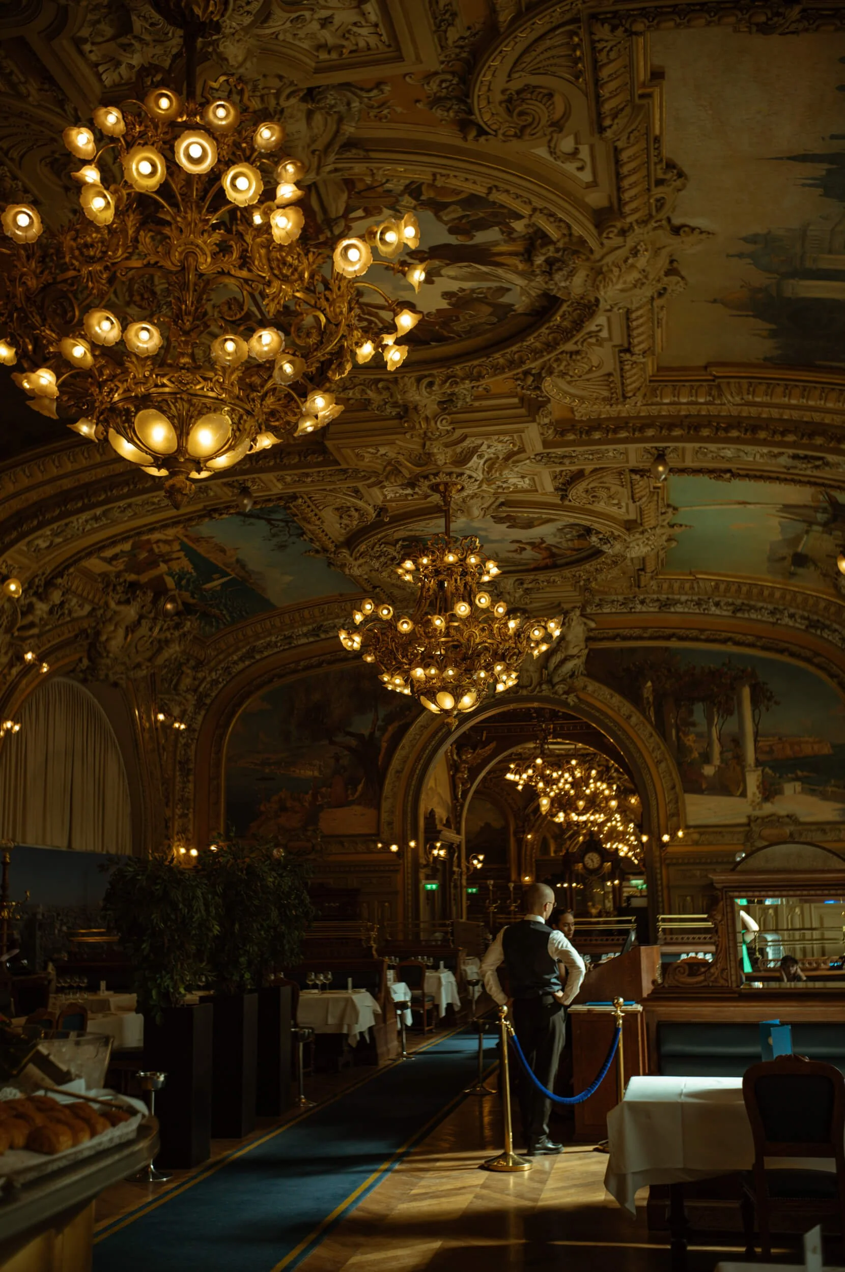 The ornate dining room of Le Train Bleu in Paris, with glowing chandeliers, painted ceilings and a waiter preparing the restaurant for breakfast.