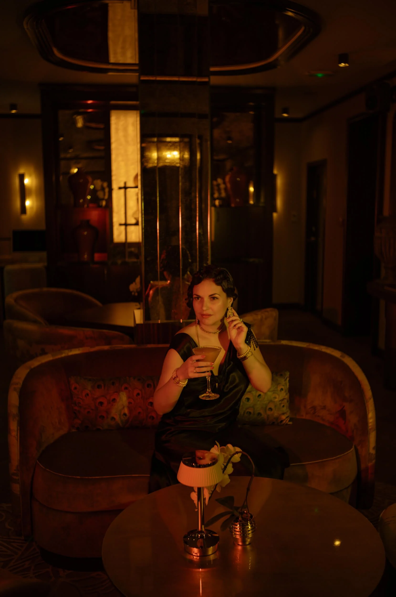 Aude reclining on a sofa at Minuit Express cocktail bar in Hôtel La Bourdonnais, Paris, photographed with warm red and amber lighting.