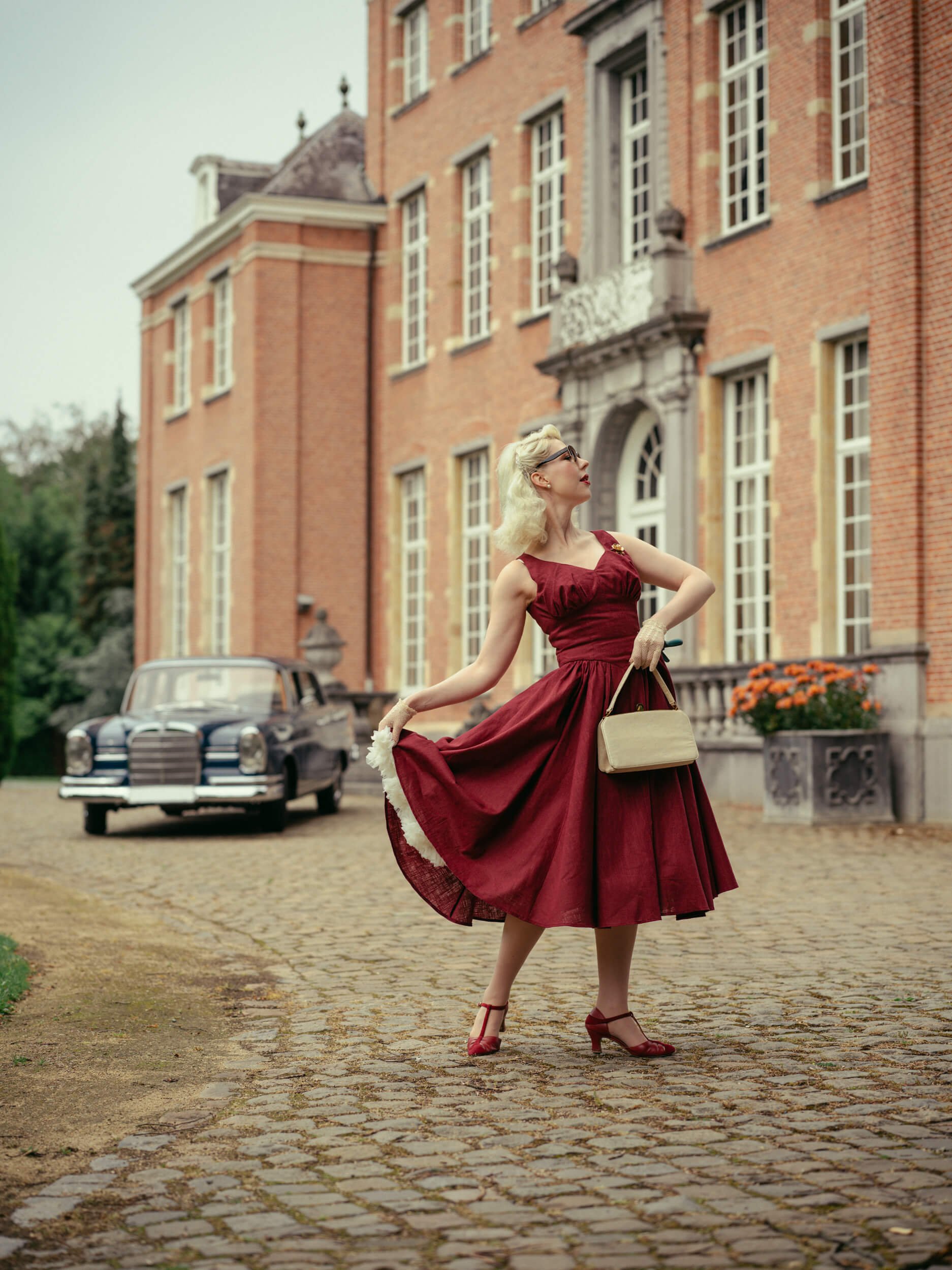 Yulia in a red dress standing beside a vintage Mercedes-Benz 220 with Château Belvedère behind her