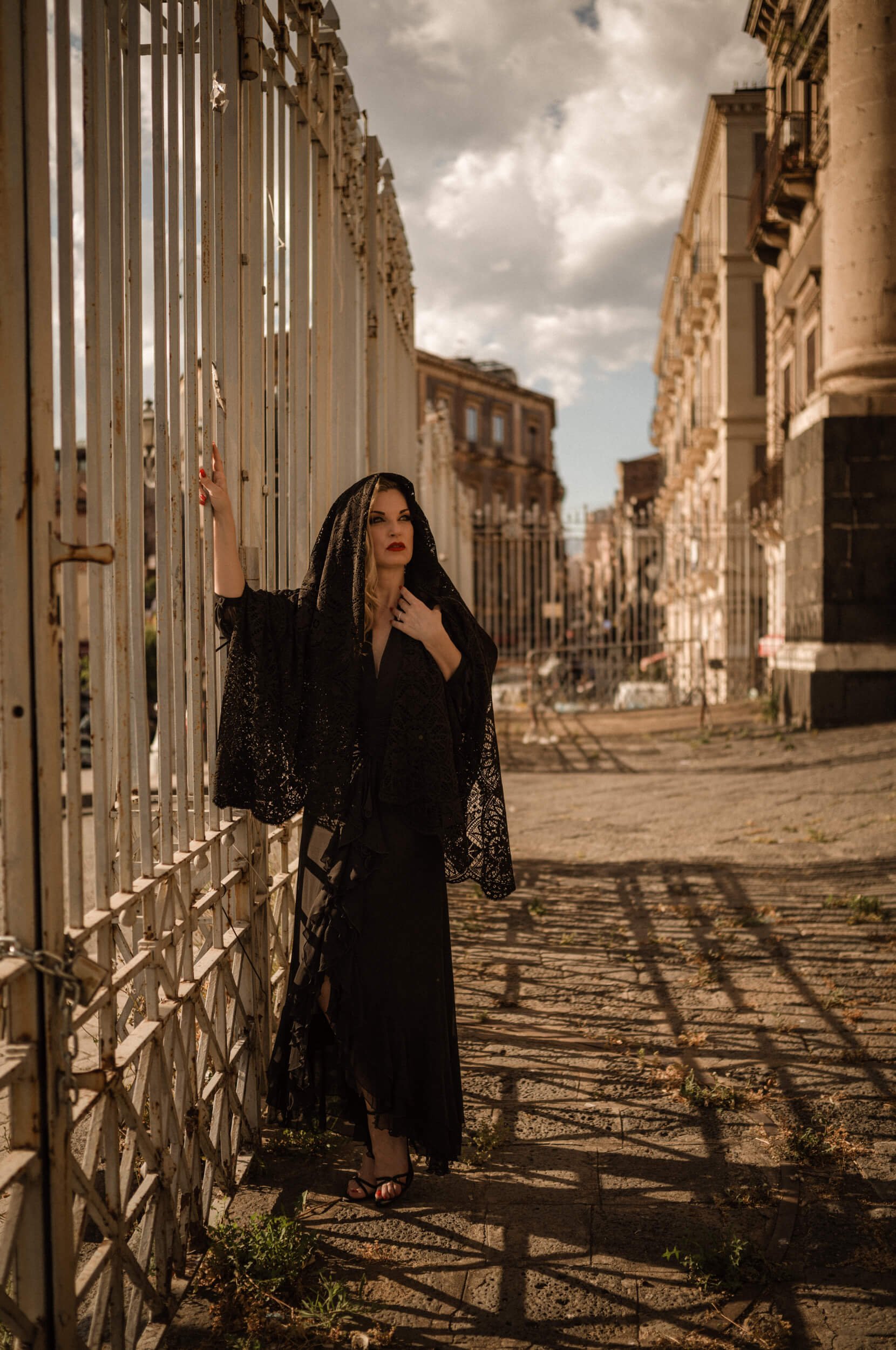 Roberta standing beside an iron gate in Catania, Sicily, wearing a black lace veil and evening dress in warm Mediterranean light.